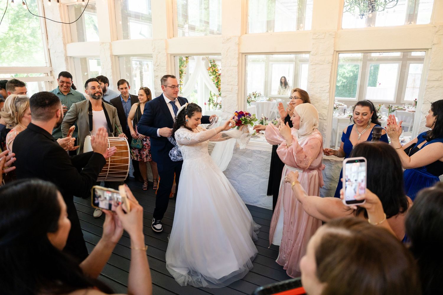 Bride and groom share first dance surrounded by cheering guests at elegant indoor wedding reception venue.