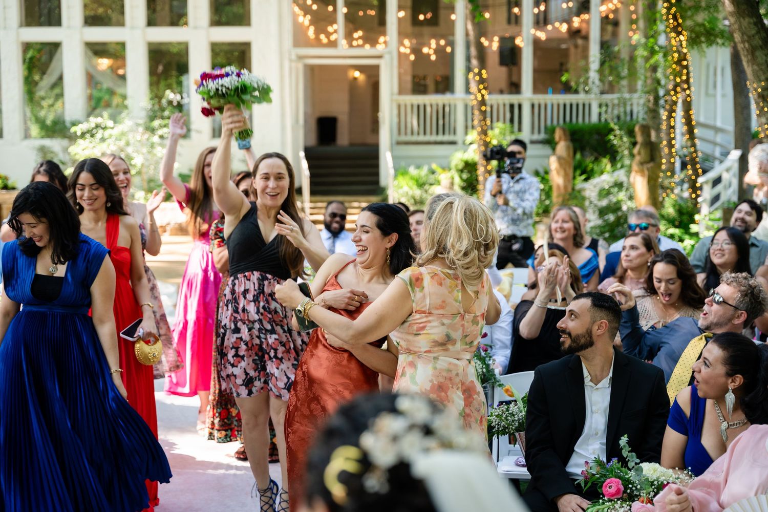 Guests celebrate at an outdoor wedding bouquet toss, with women laughing and embracing on a floral-decorated venue aisle.