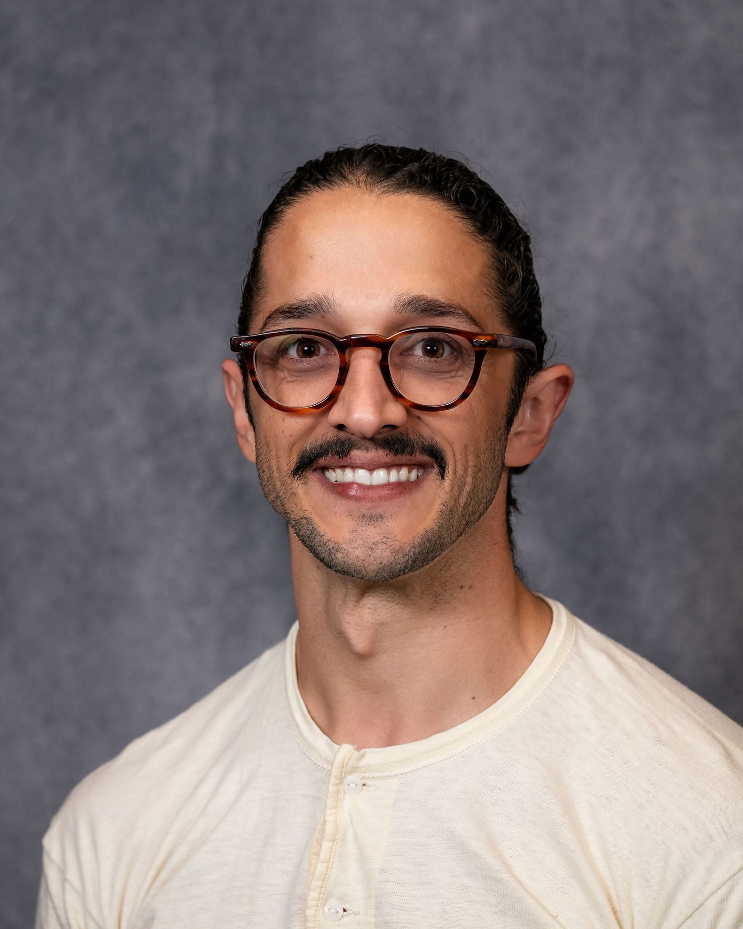 Smiling man with glasses, mustache, and pulled-back hair wearing a white henley shirt against a gray background.