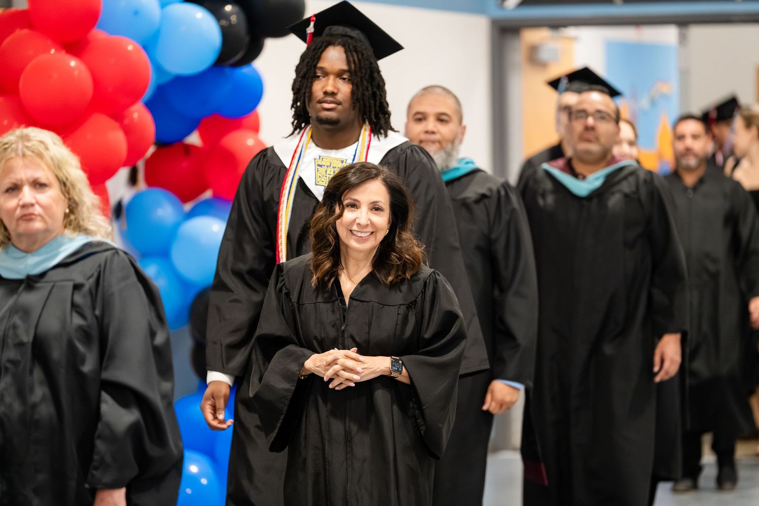 Graduates in black caps and gowns walk in procession at a graduation ceremony with colorful balloons in the background.