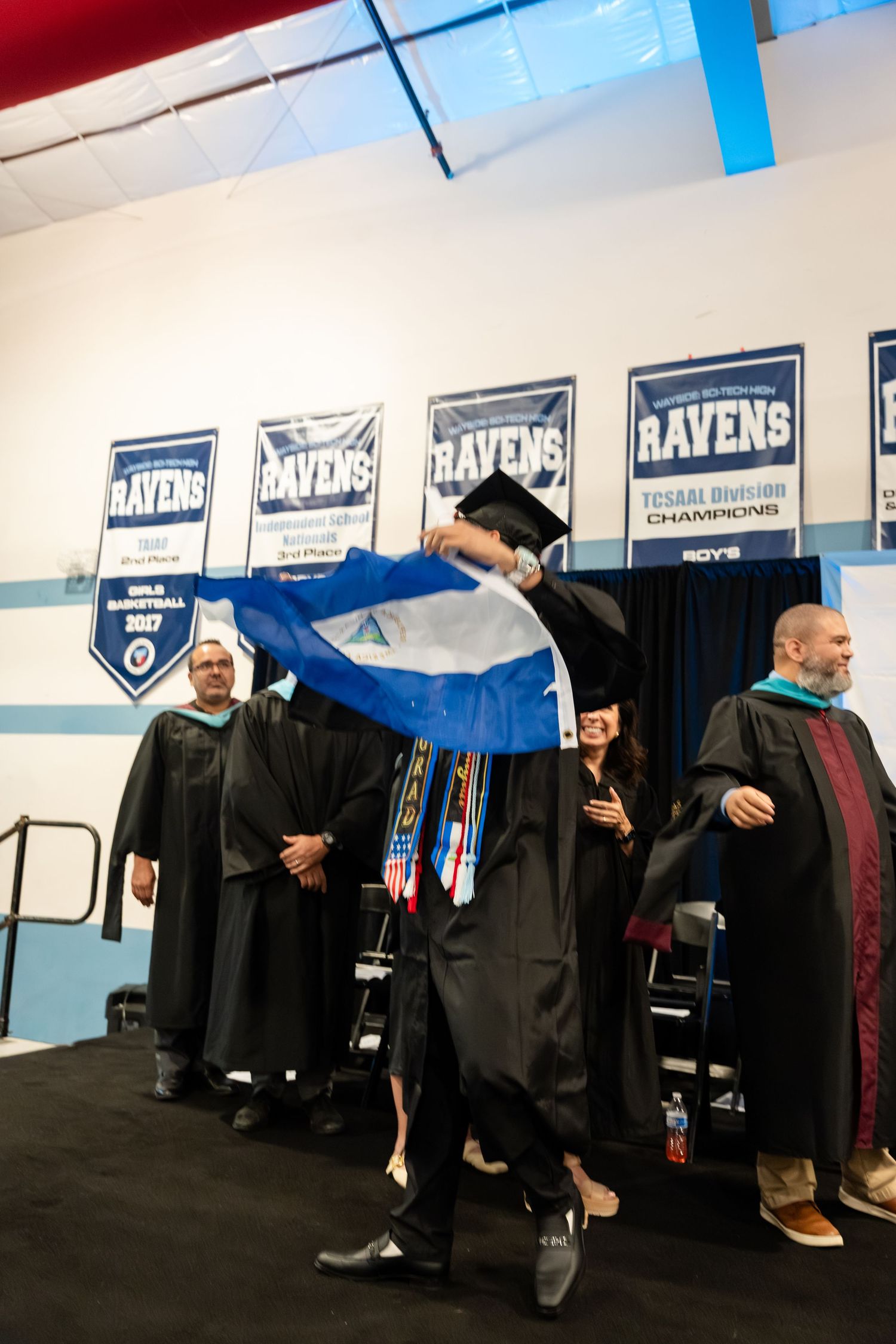 Graduate waves Nicaragua flag proudly during Ravens college graduation ceremony in academic regalia.
