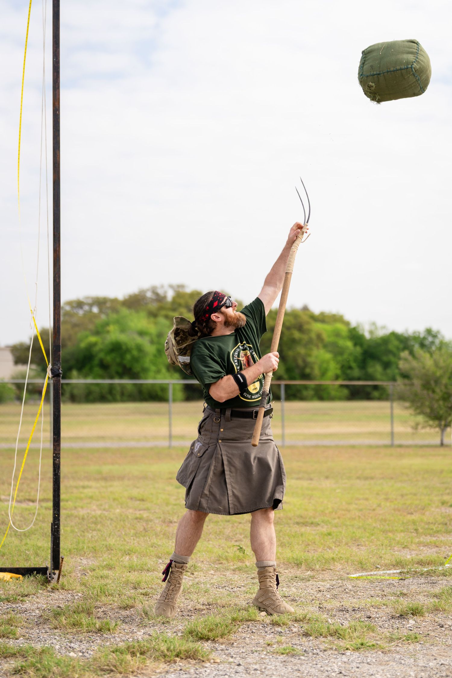 Person tossing a green bag in an outdoor field, wearing a kilt, boots, and green shirt during a Highland Games competition.