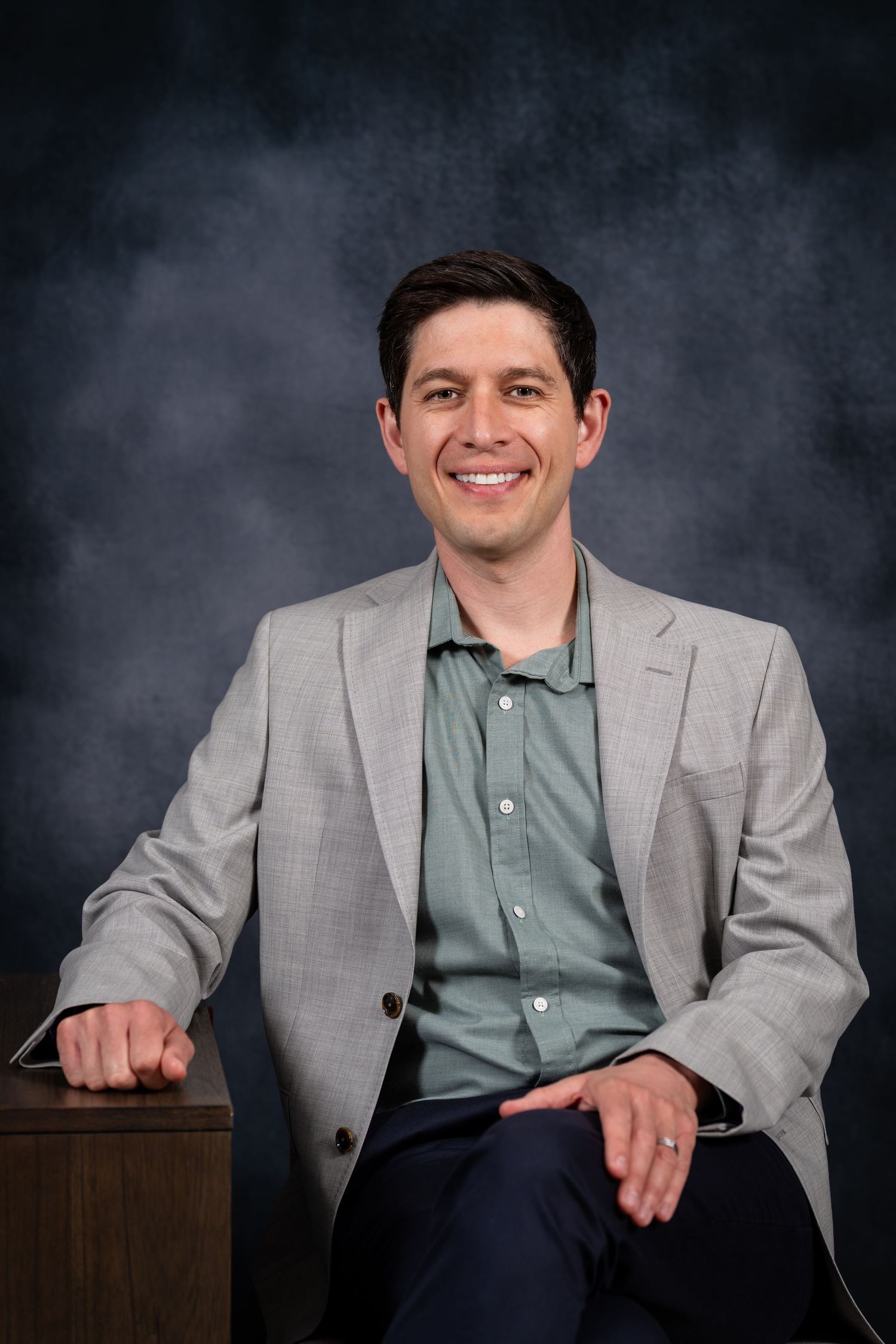 Smiling man in gray blazer and green shirt seated casually against a dark studio background.