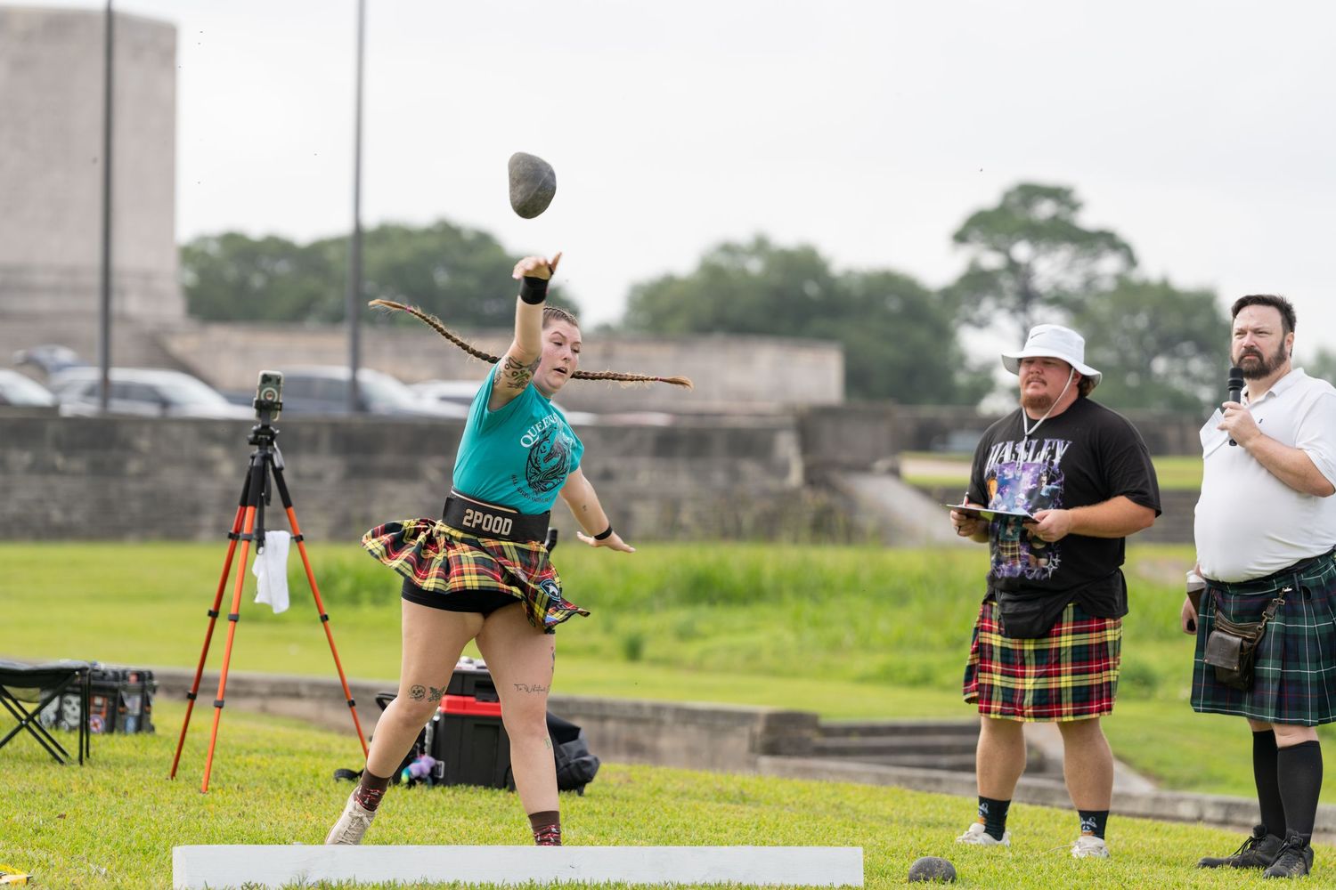 Woman competing in Highland Games throwing event, wearing tartan skirt, with judge in kilt watching outdoors.