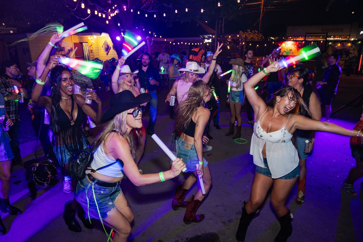 People dancing at an outdoor night festival with colorful glow sticks and neon lights.