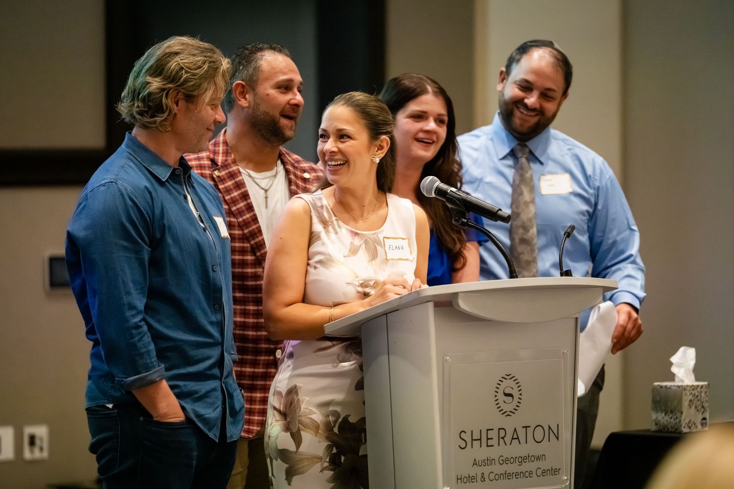Group of five people laughing together at a Sheraton Austin Georgetown Hotel podium during a conference event.