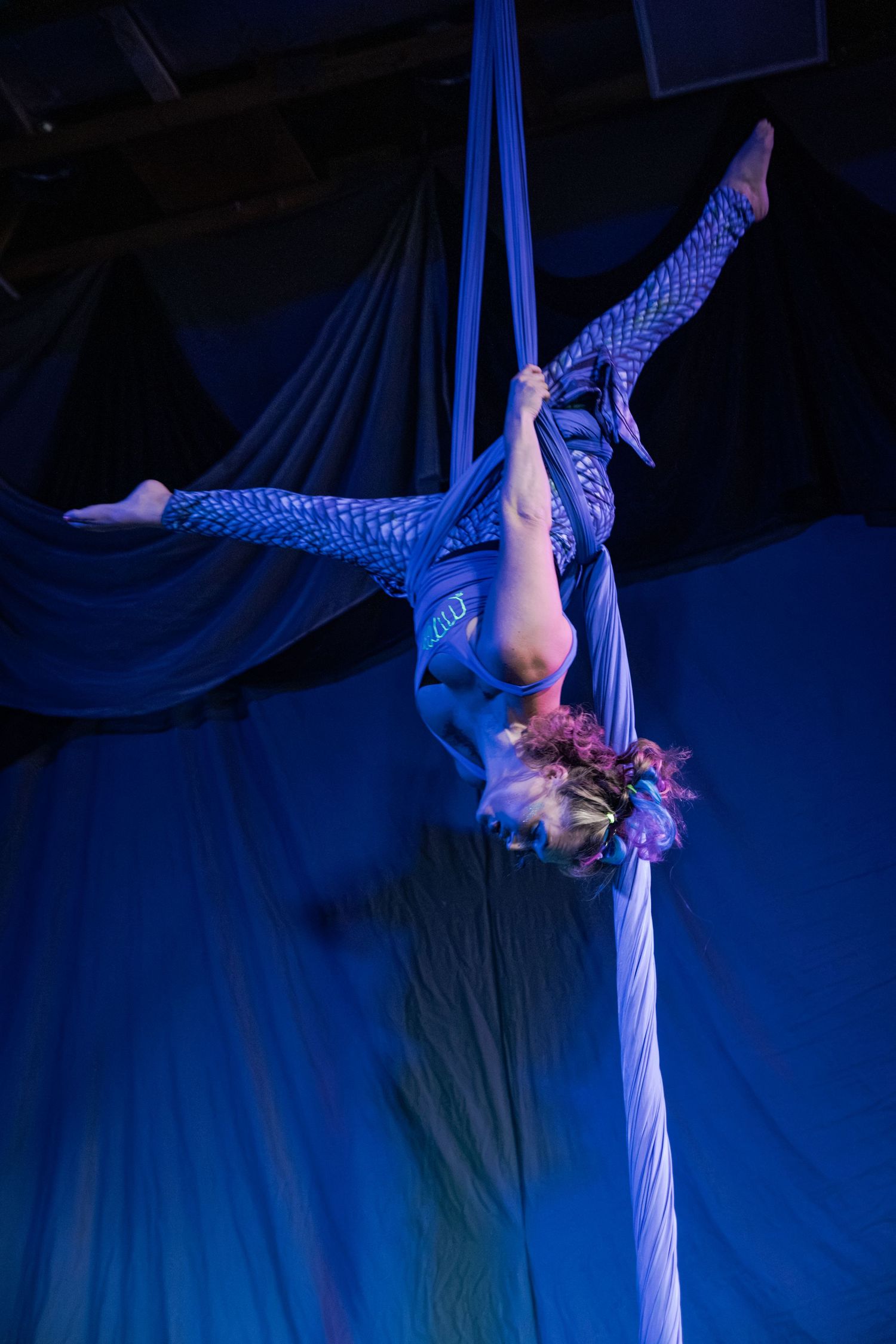 Aerial silk performer in blue costume suspended upside down on blue fabric drapes under dramatic stage lighting.