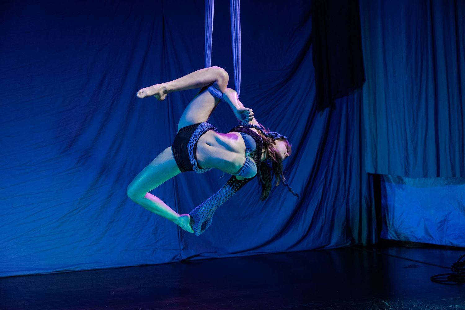 Aerial silk performer executing a dramatic backbend pose suspended mid-air against a blue-lit stage backdrop.