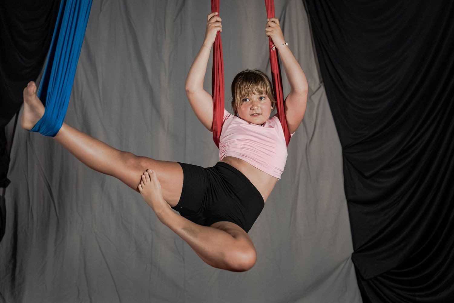 Young gymnast performing aerial silk routine, suspended mid-air in splits pose with red and blue fabric.