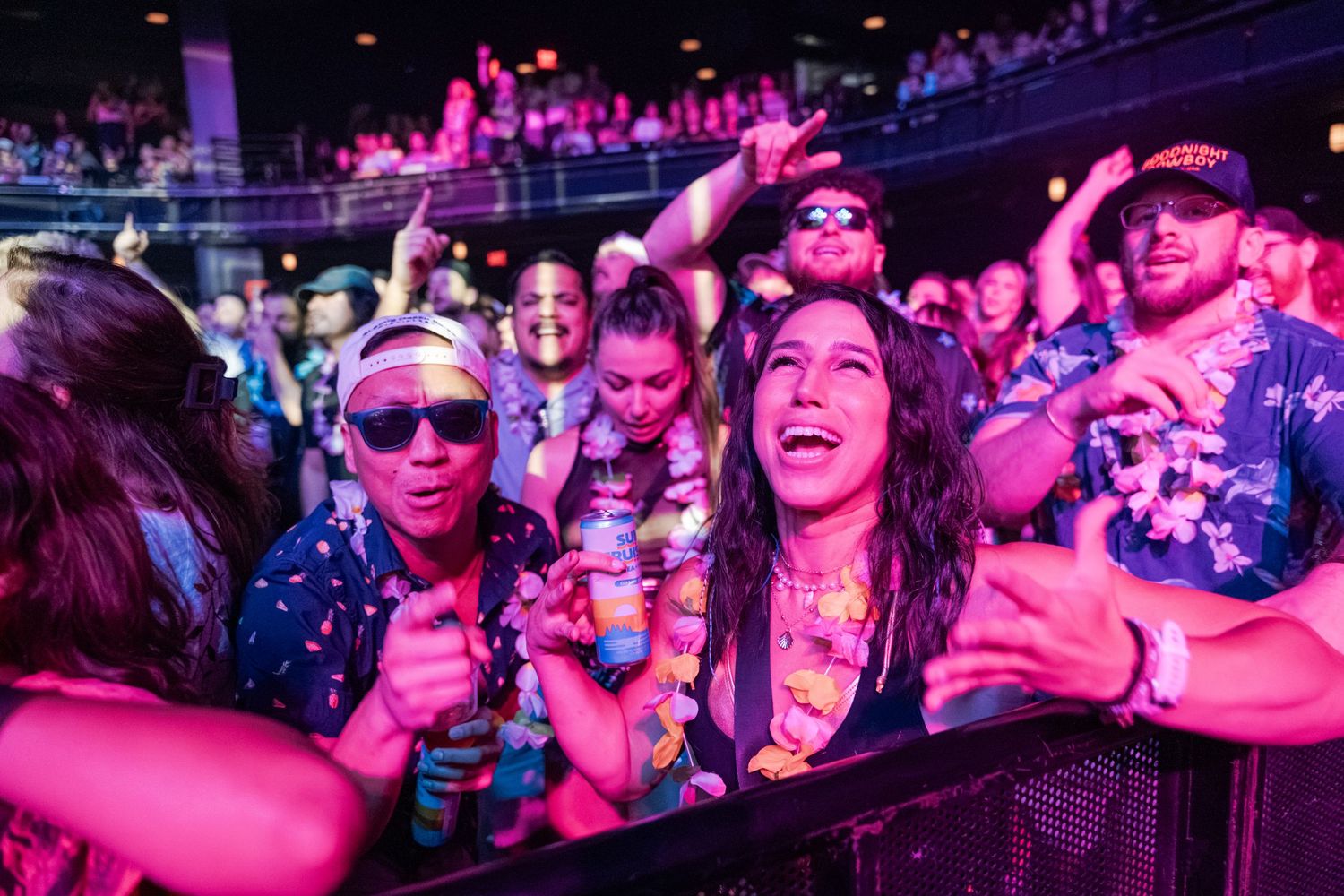 Excited crowd enjoying a live concert under pink and purple lights, dancing and cheering at an indoor music venue.