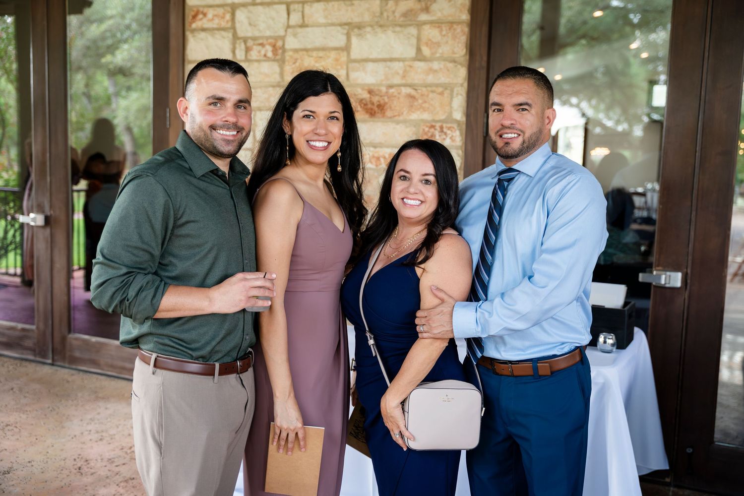Group of people in formal attire posing together at a social gathering with stone wall backdrop.