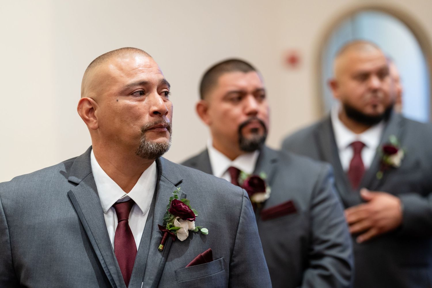 Groomsmen wearing burgundy ties and grey suits standing at altar during wedding ceremony.