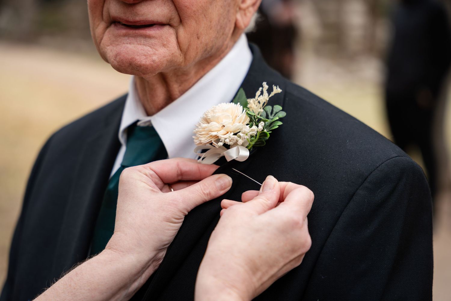 Someone's hands pin a delicate cream and white floral boutonniere onto a dark suit jacket with teal tie.