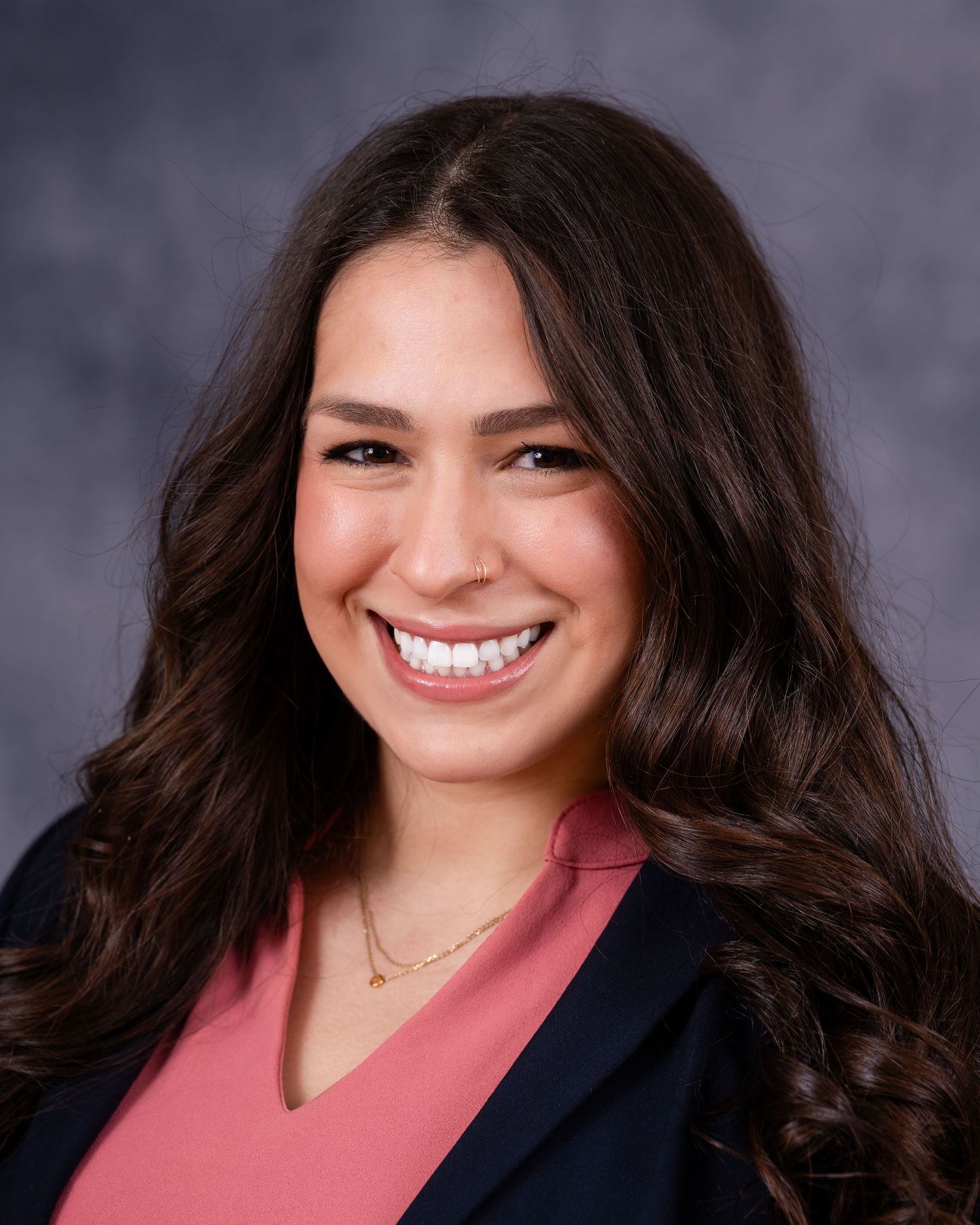 Professional portrait of a smiling person wearing a pink blouse and navy blazer against gray backdrop.