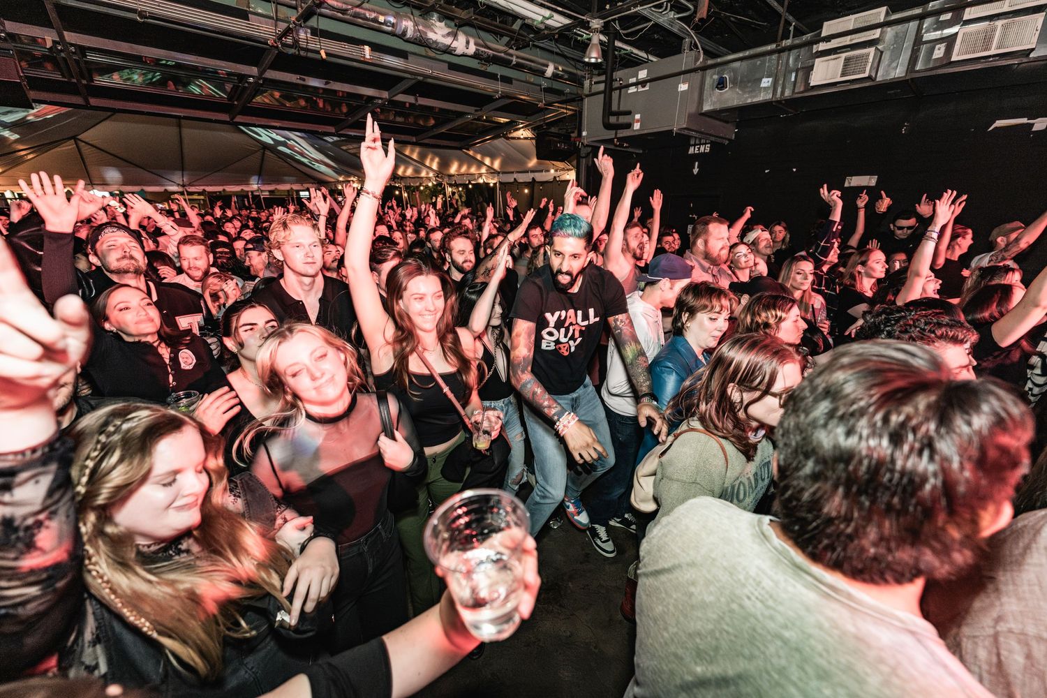 An energetic crowd raises their hands and drinks at a packed concert venue with red lighting overhead.