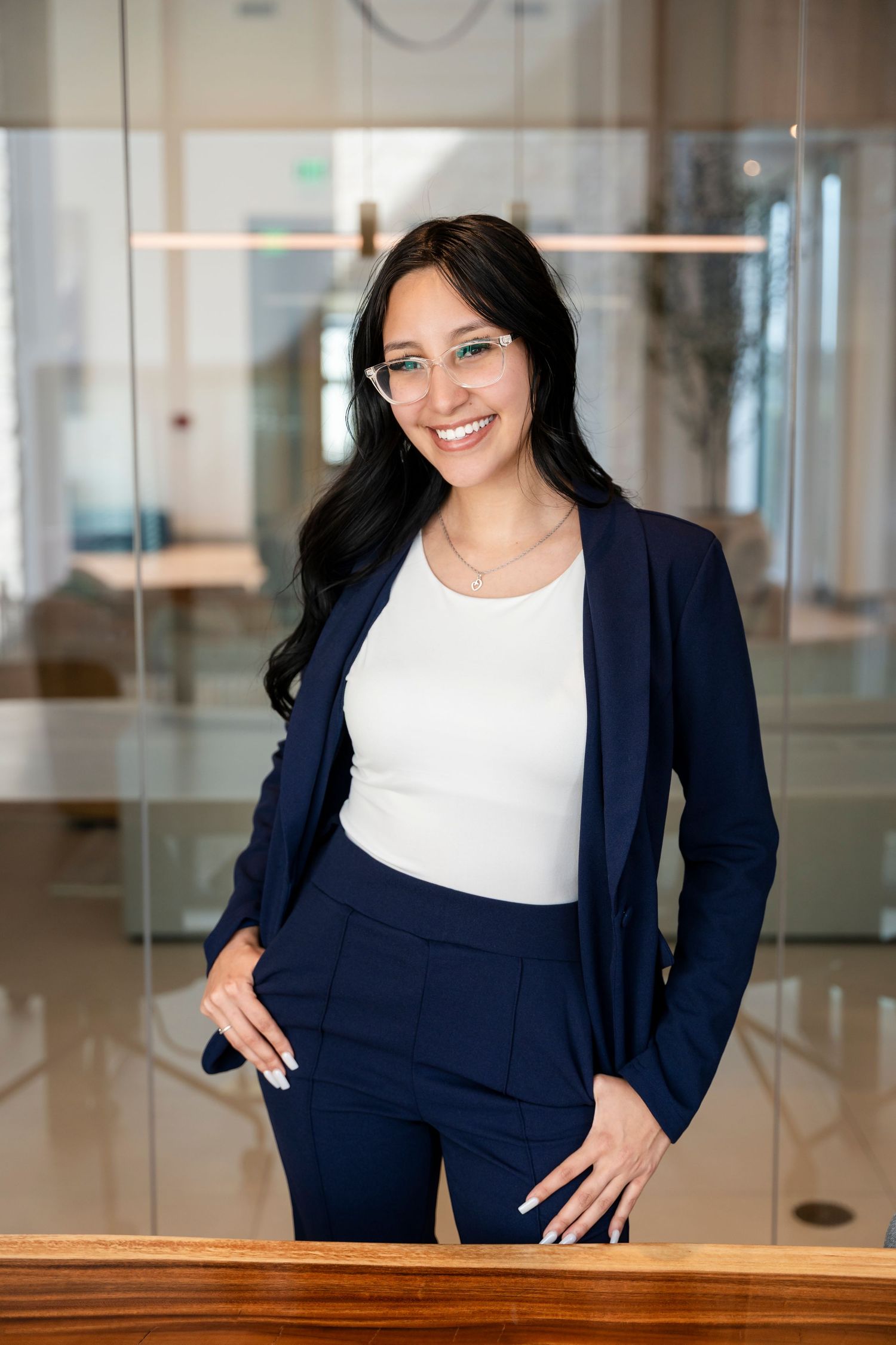 Professional wearing a navy blue business suit and glasses stands confidently in an office environment.