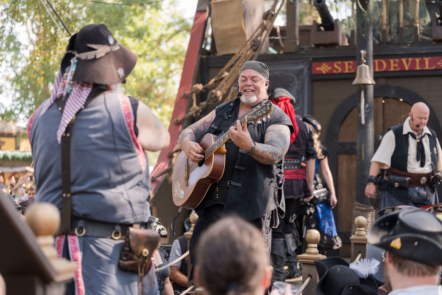 Musicians perform on stage at a renaissance festival with acoustic instruments and period costumes.