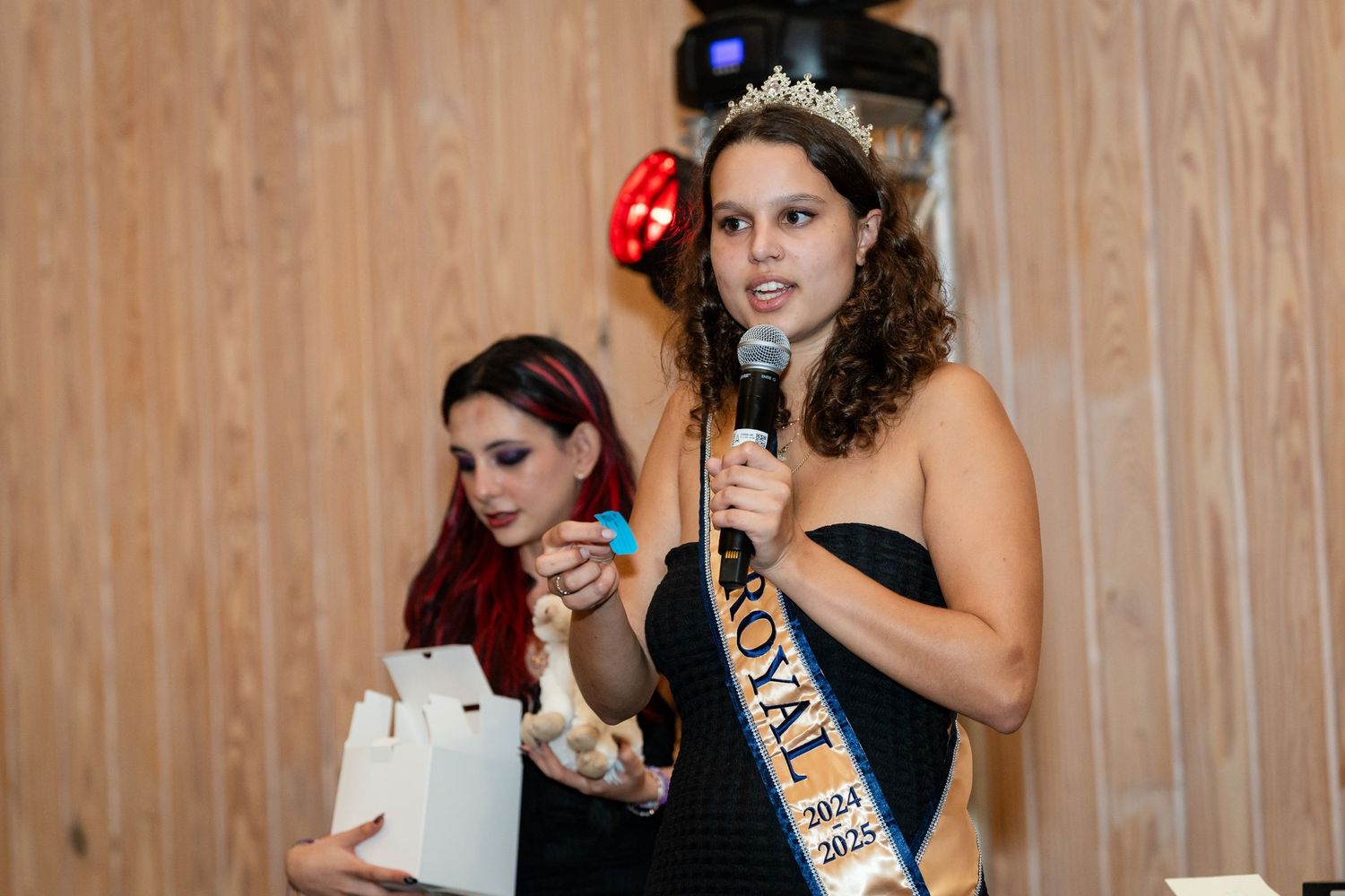 Two people speaking into microphones at an indoor event with wooden walls.