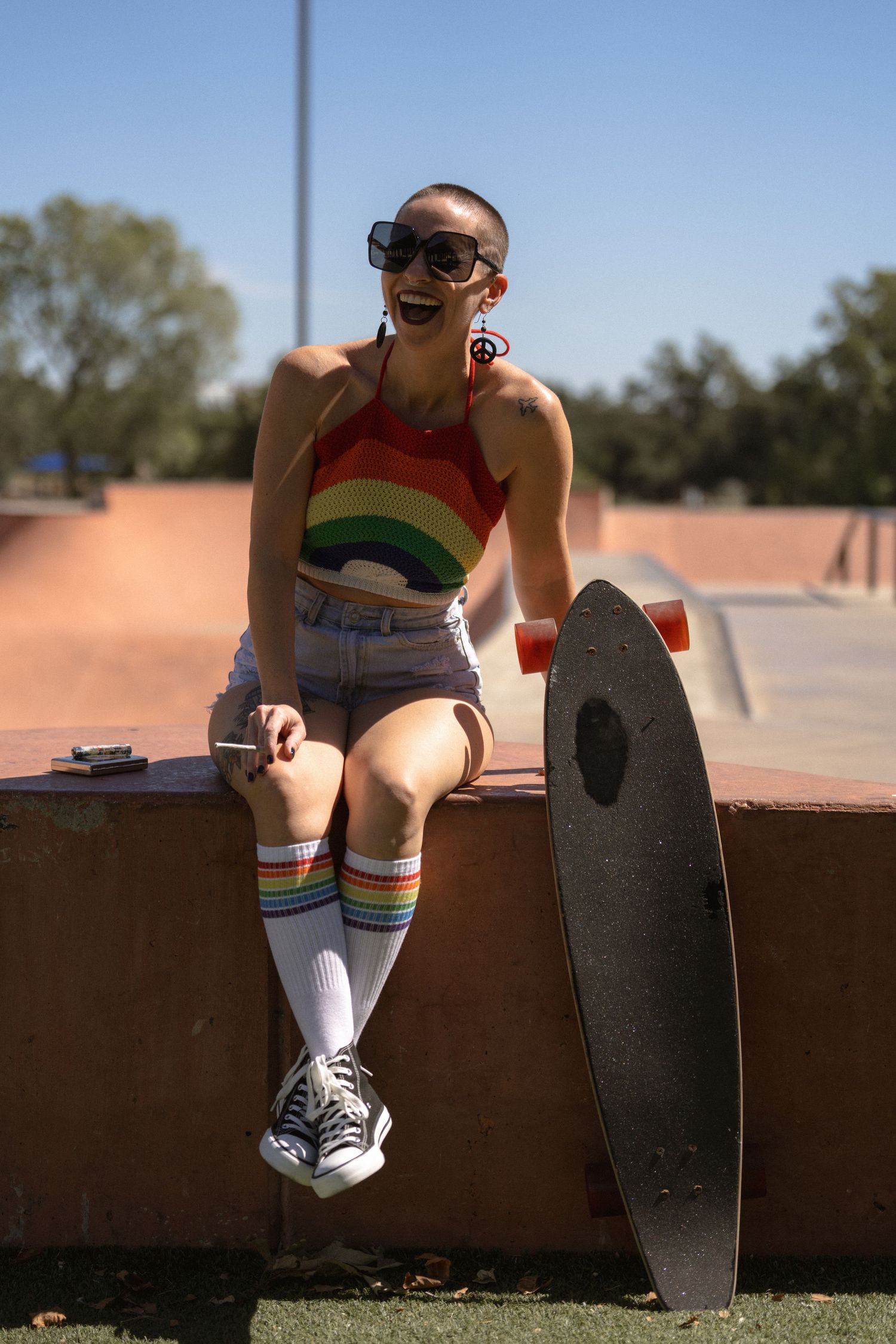 Skater in rainbow tank top and striped socks sits with skateboard at skate park.