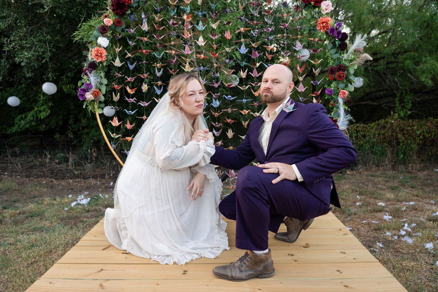 A couple in wedding attire sits on a wooden platform under a colorful string backdrop during an outdoor ceremony.