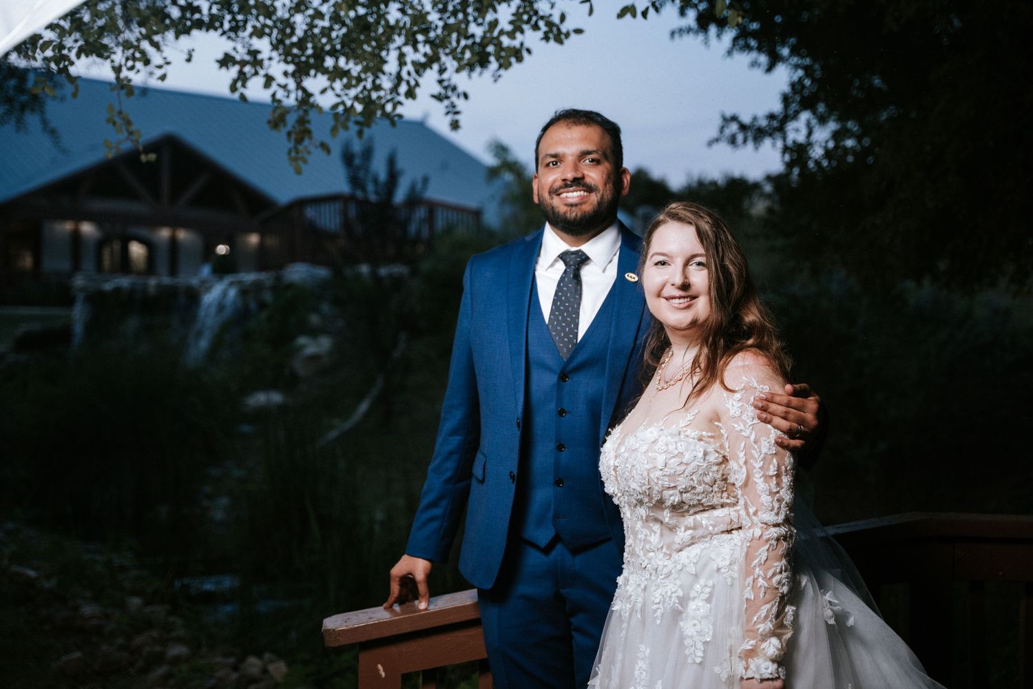 A couple poses outdoors at dusk in formal wedding attire near a building.