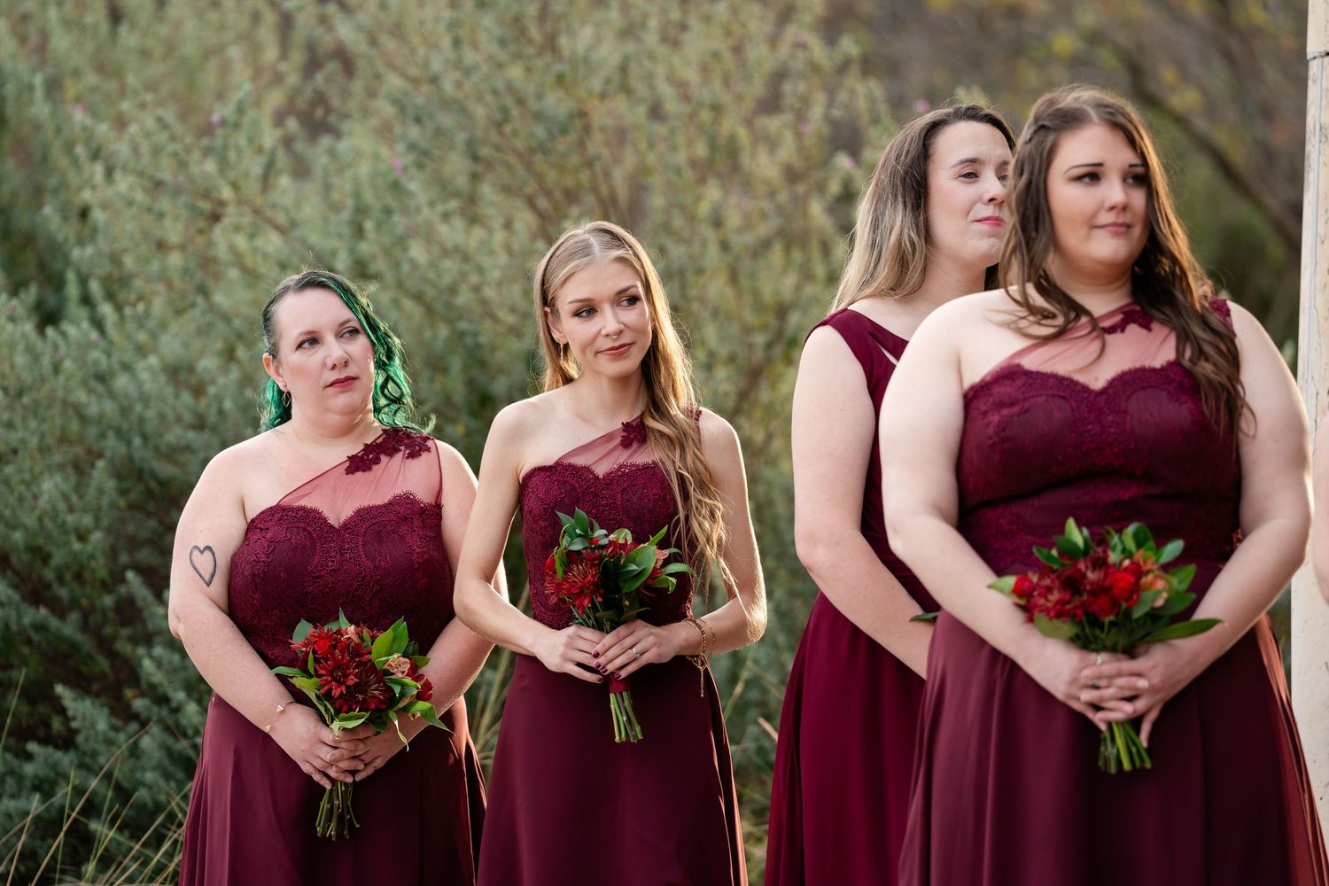 Bridesmaids in burgundy dresses hold red bouquets while standing outdoors at a wedding ceremony.