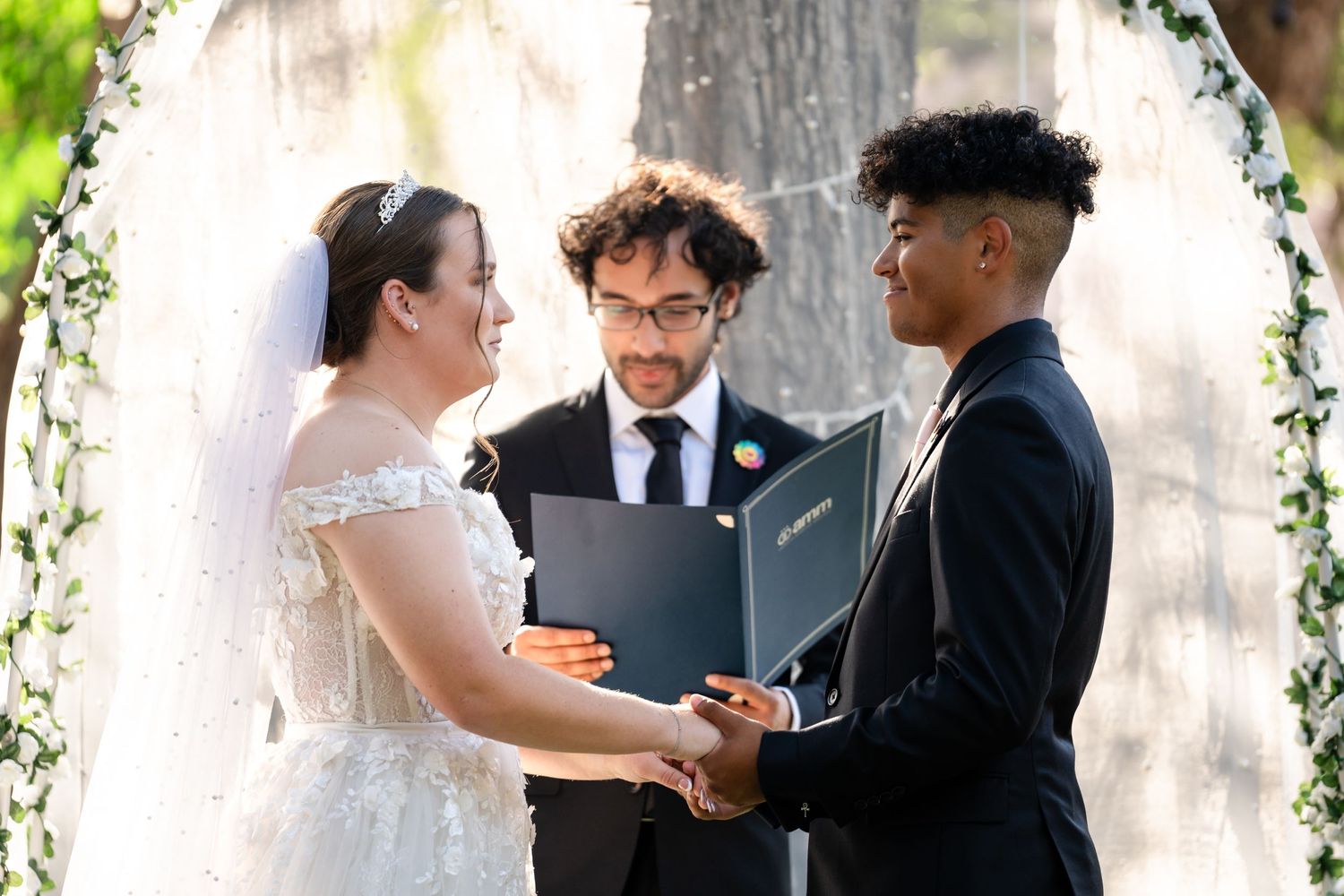 A wedding ceremony takes place under a floral arch with the officiant conducting the service outdoors.