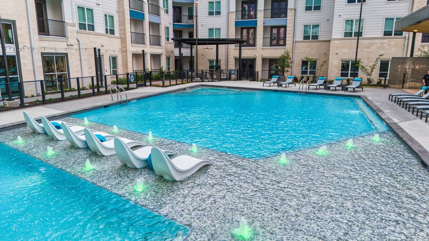 Resort-style pool with in-water lounge chairs and modern apartment buildings in background.
