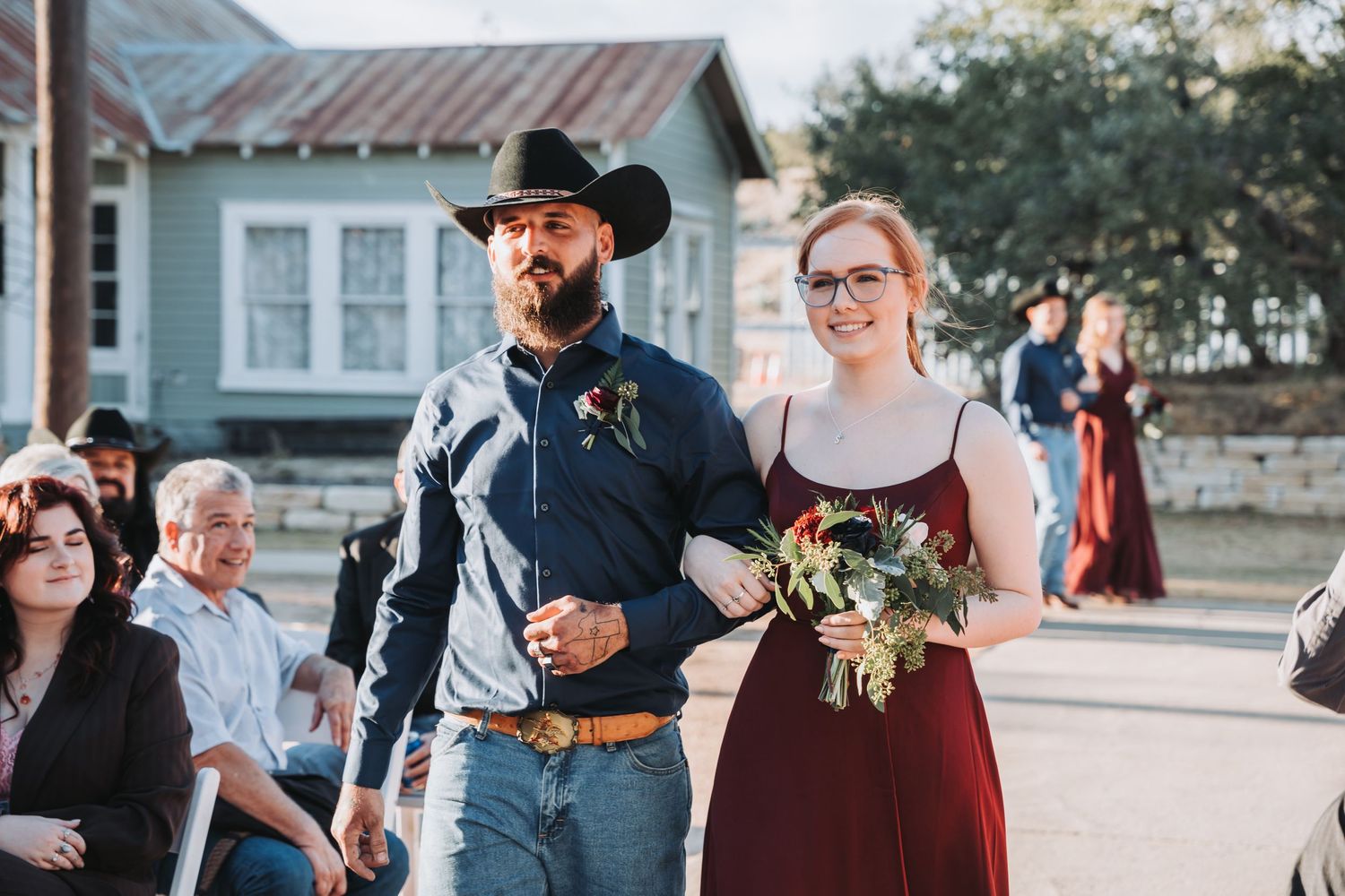 A couple walks arm in arm during an outdoor ceremony while holding a floral bouquet.