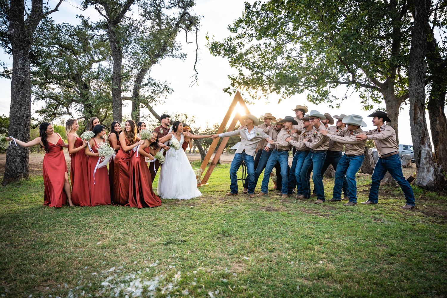 Wedding party in red dresses and blue jeans strikes playful pose on grassy lawn at outdoor venue.