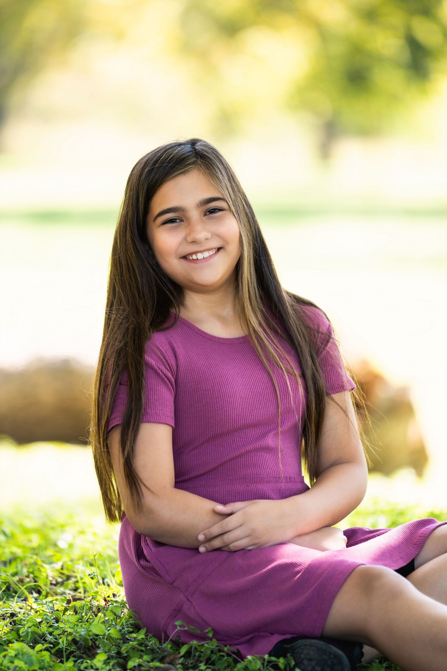 Person wearing purple dress sits on grass with gentle smile in outdoor portrait setting.