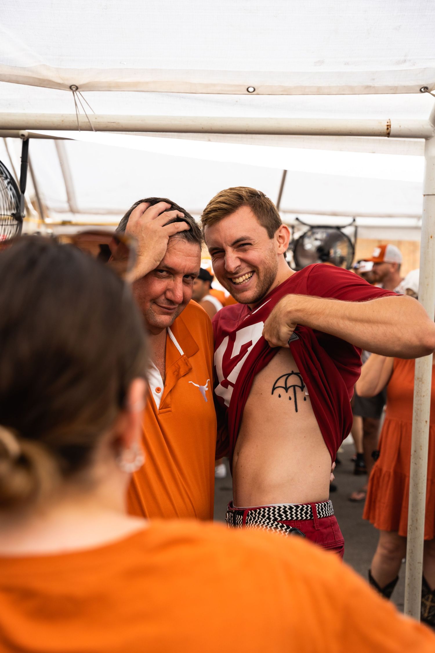 A group of people in orange and red shirts gather under a white tent at an outdoor event.
