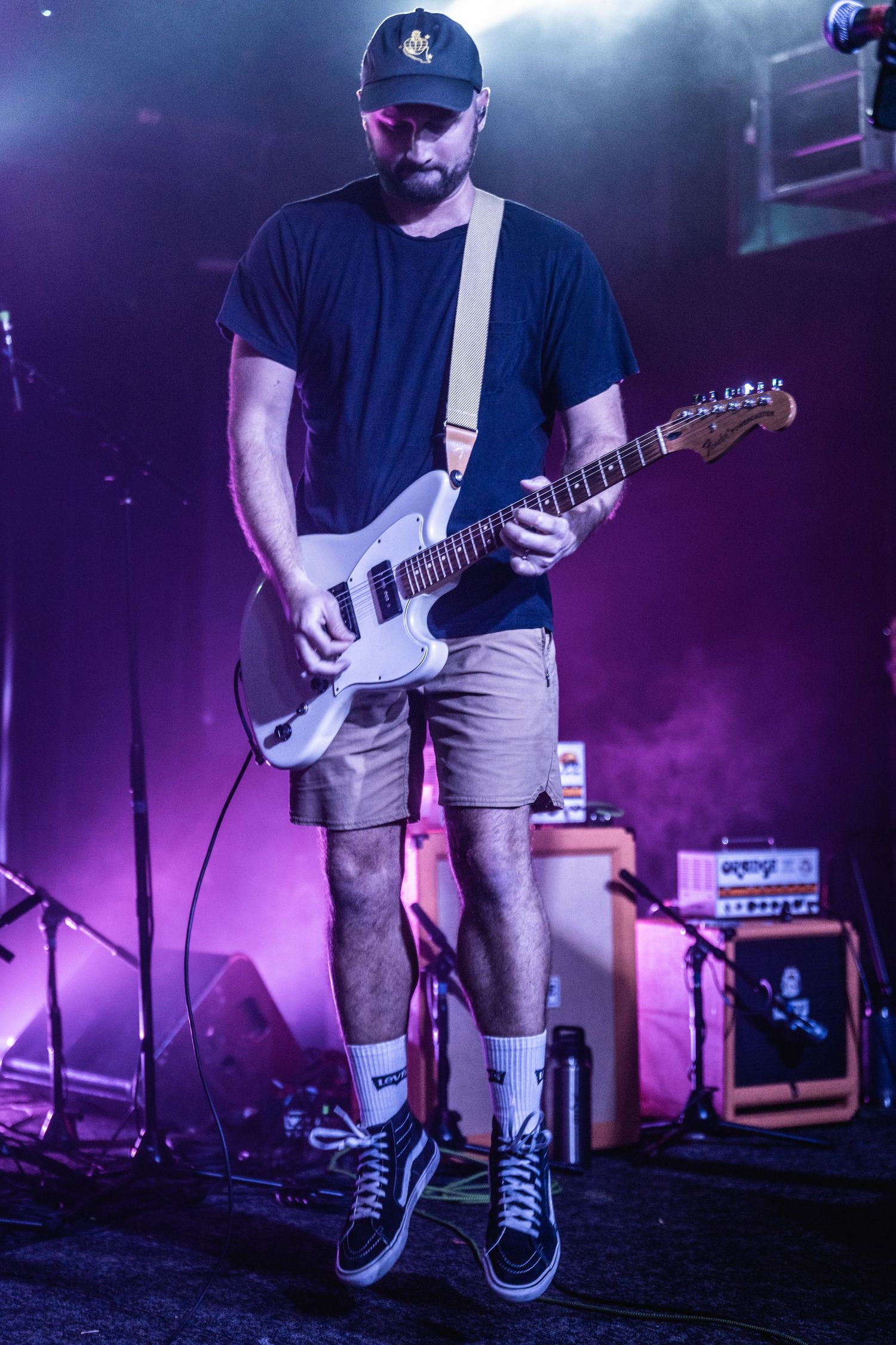 Musician plays electric guitar on stage with purple atmospheric lighting and amp setup in background.