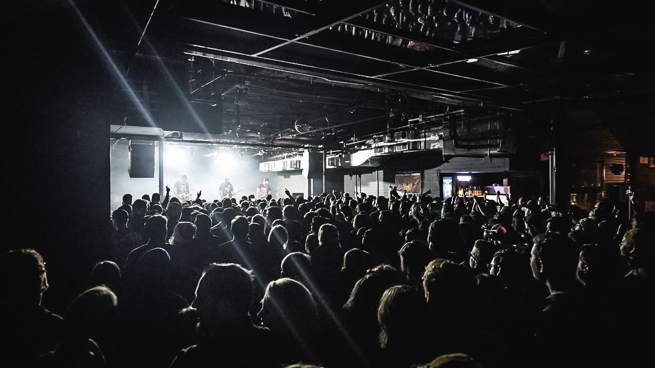 Silhouetted crowd of concert attendees in a dark venue with dramatic lighting from the stage.
