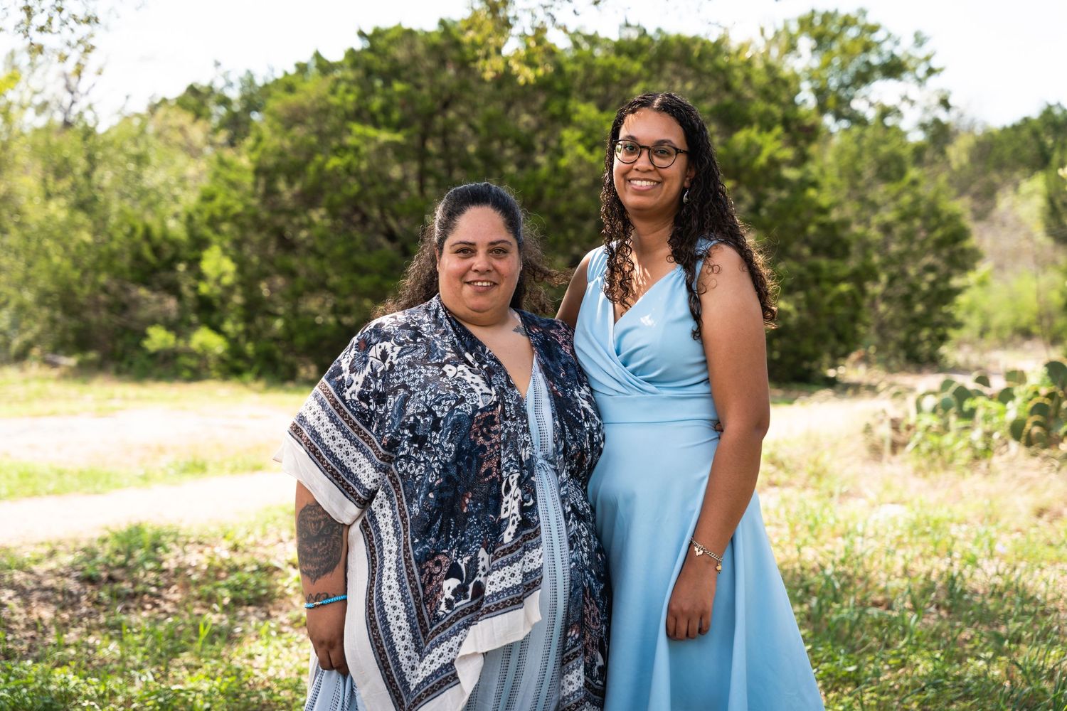 Two people wearing blue dress and patterned dress pose together in outdoor setting.