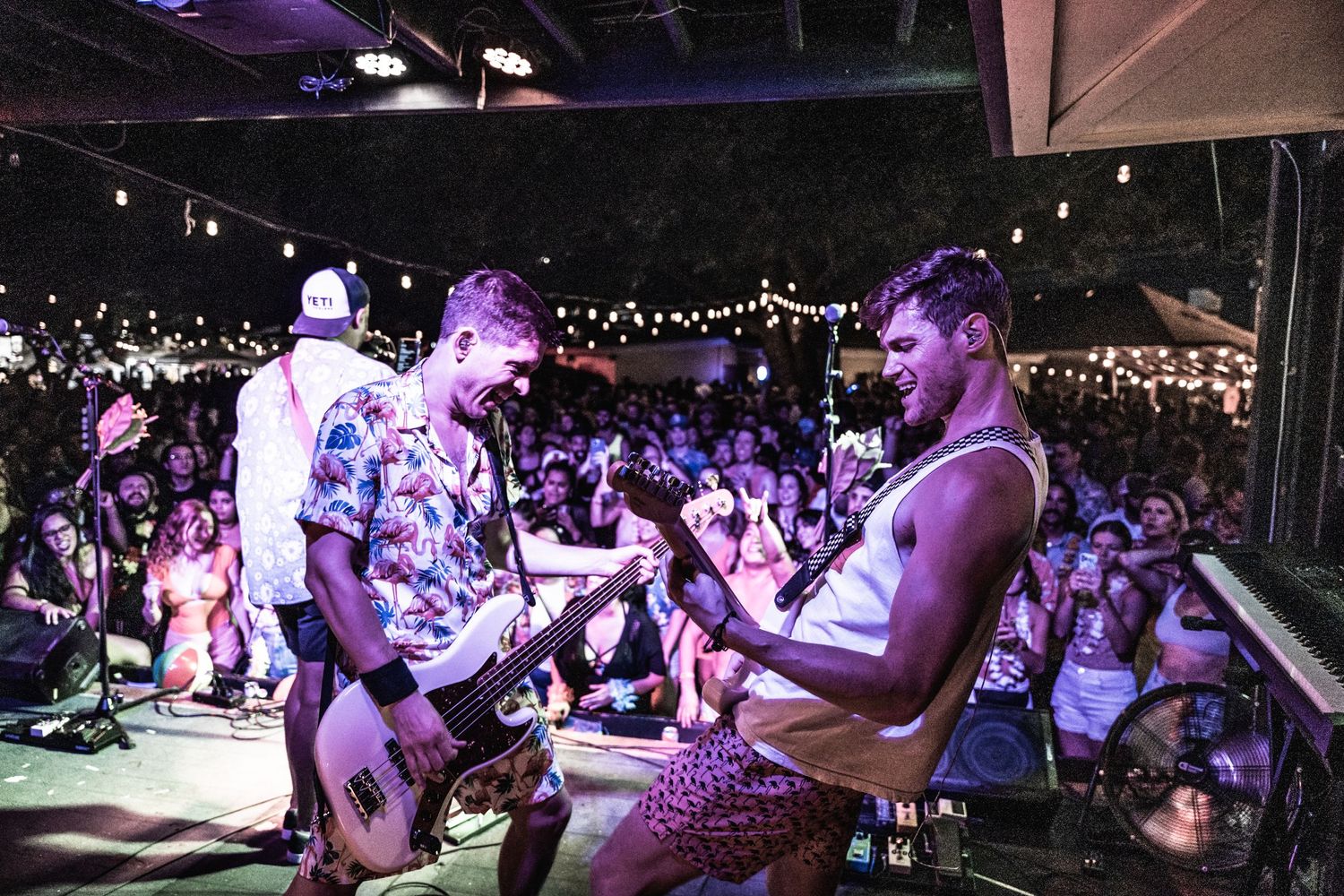 Musicians perform energetically on stage at a crowded concert venue with purple lighting.