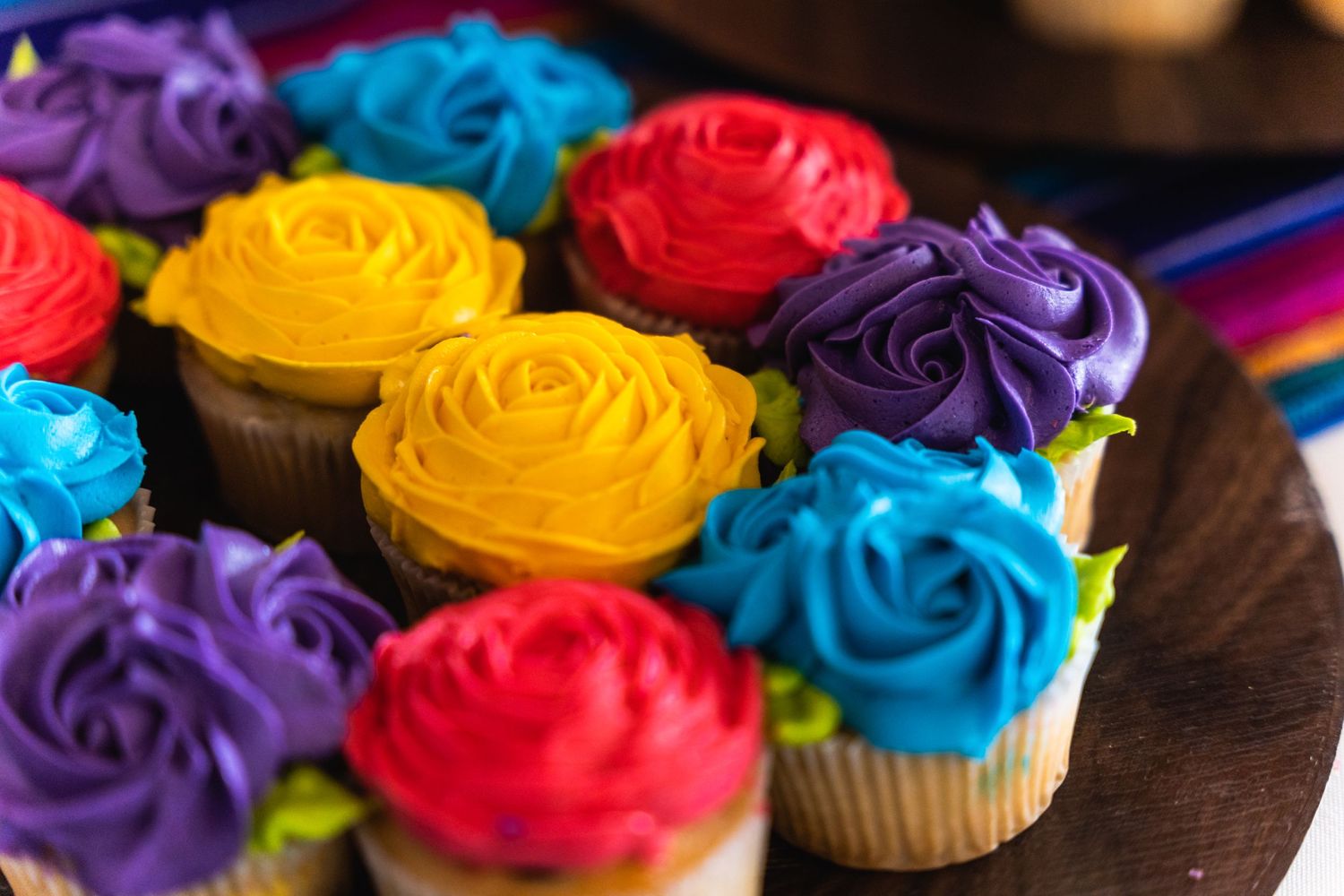 Colorful rose-decorated cupcakes in purple, yellow, red and blue frosting arranged on a dark plate.