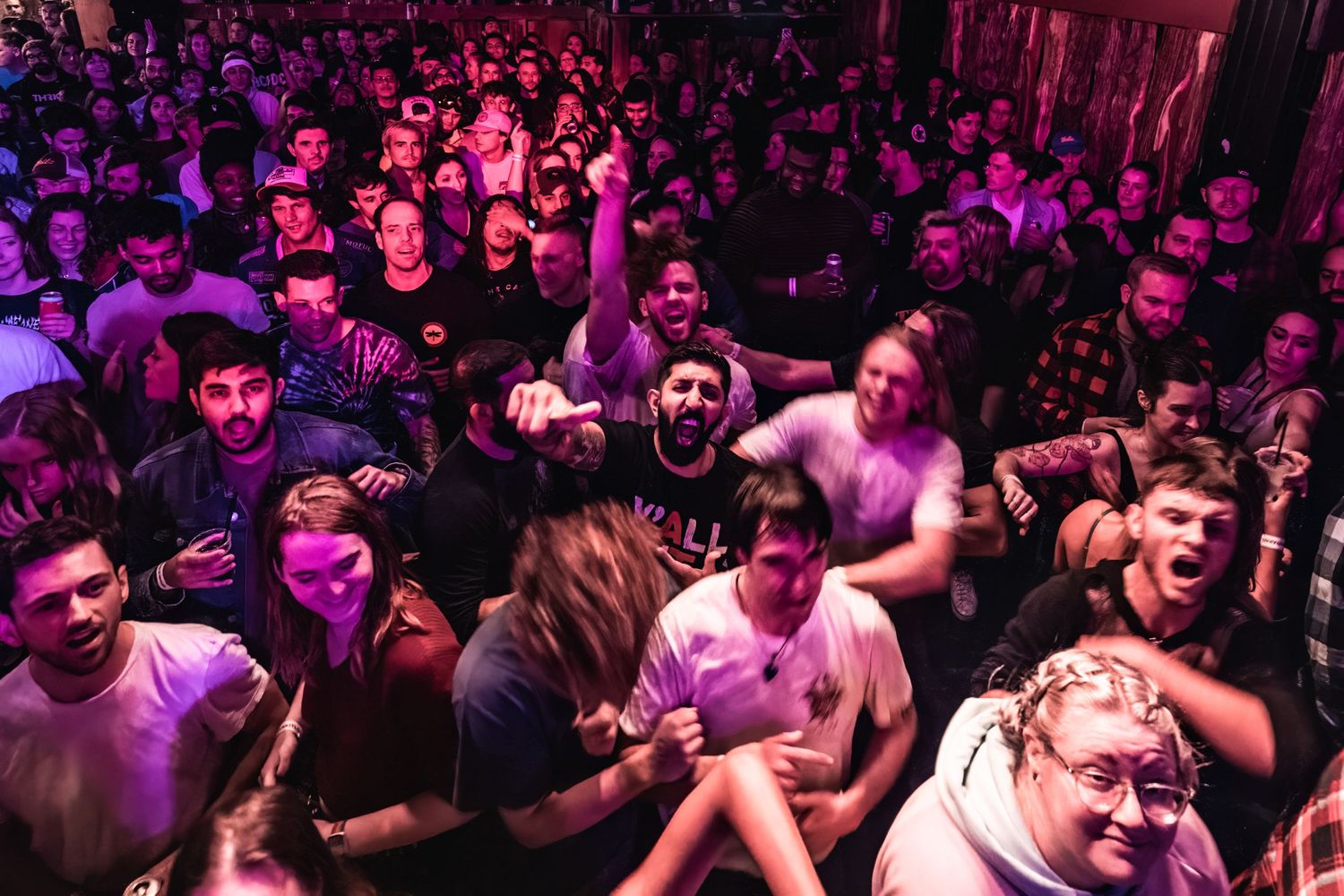 Crowded concert venue with energetic audience illuminated by purple lighting during live performance.