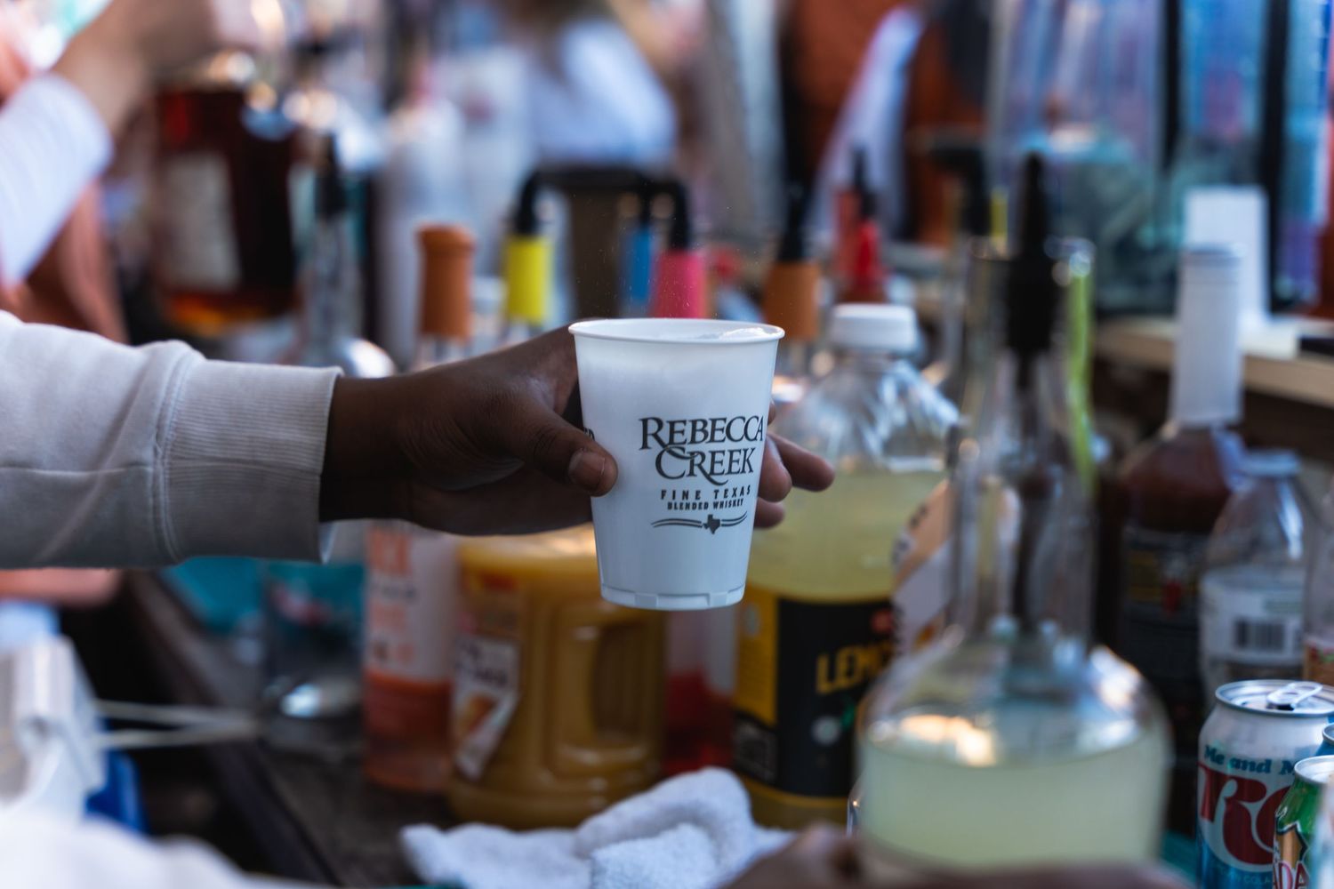 A hand holds a branded disposable cup at a bustling beverage service station.