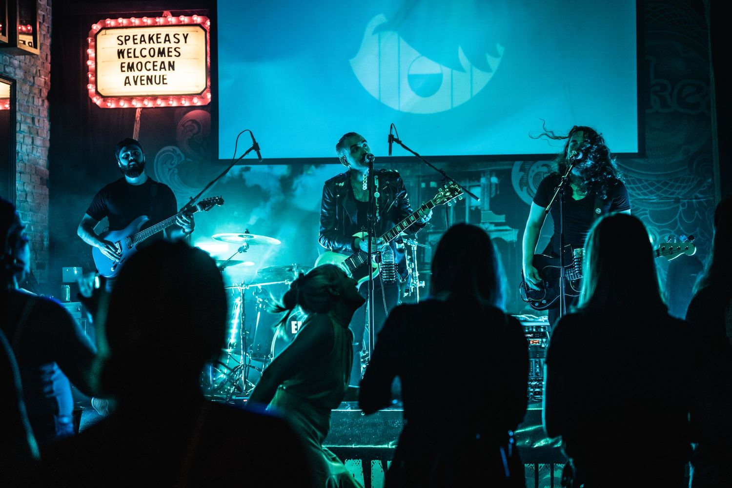 Band performing on stage with blue lighting and silhouetted audience in foreground.