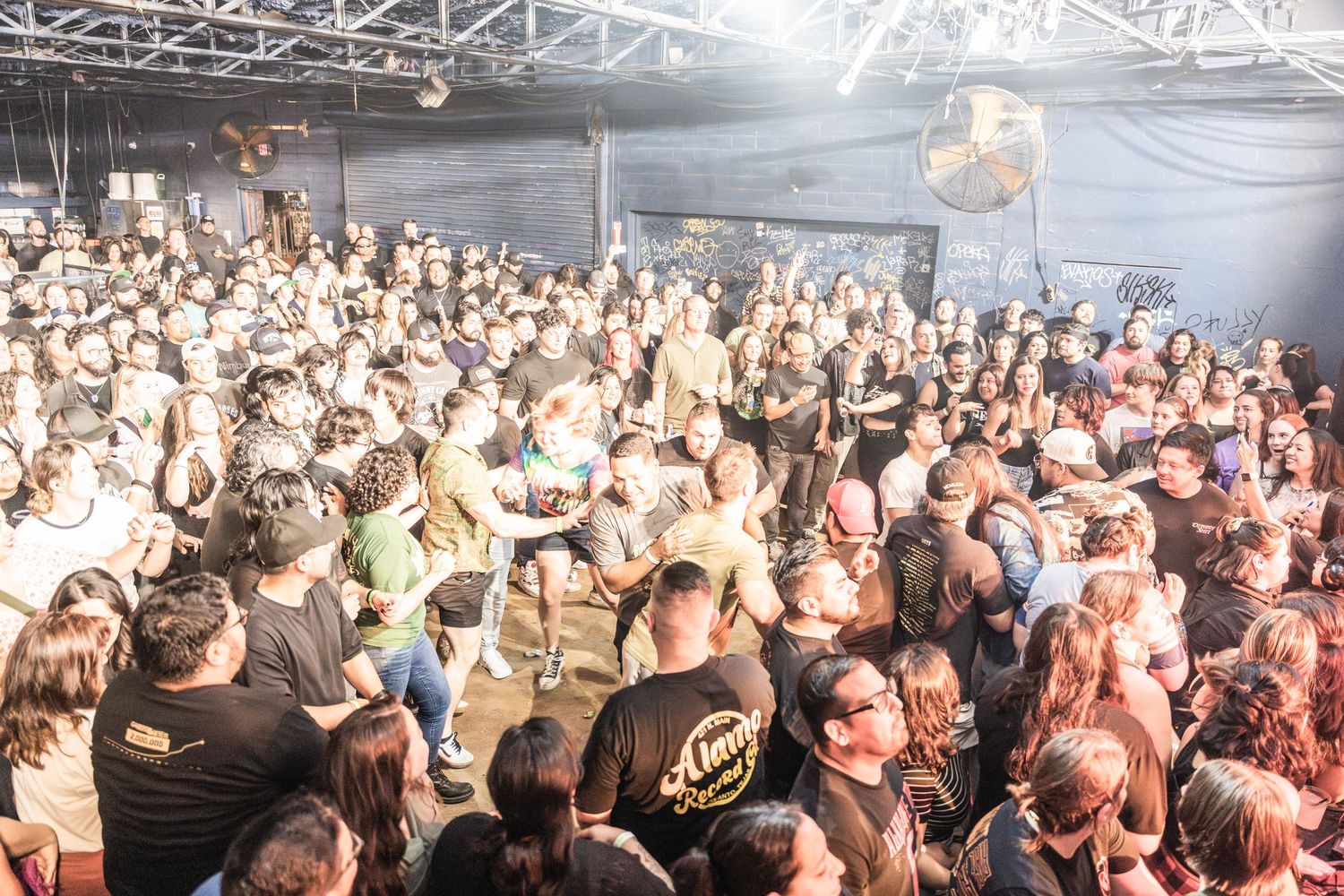 Large crowd of concertgoers packed tightly together at an indoor music venue with industrial ceiling and lighting.