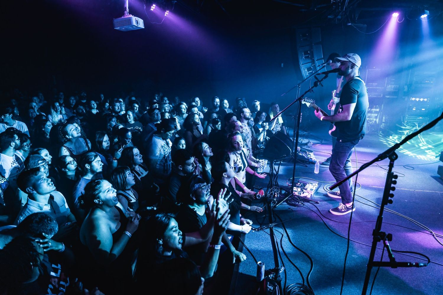 Concert crowd watches an energetic live performance with purple and blue stage lighting.