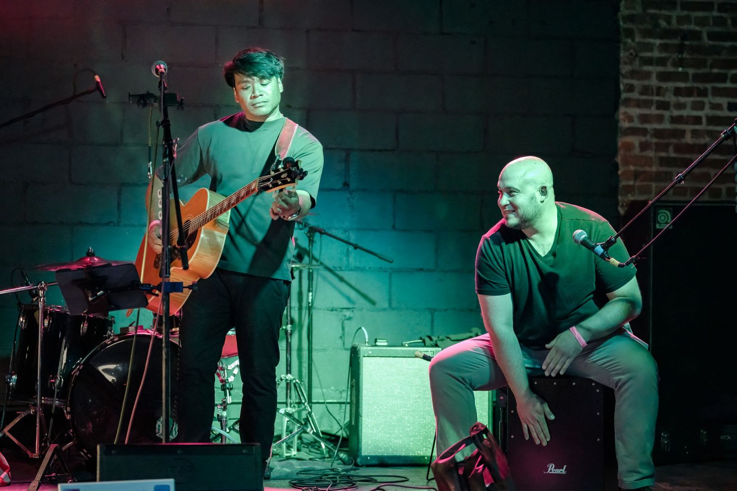Two musicians perform on stage with acoustic guitar and percussion setup under teal lighting.