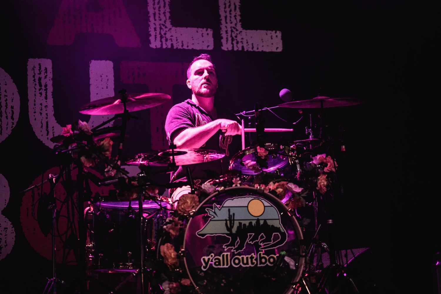 Drummer performs behind drum kit on dimly lit stage with purple lighting effects.