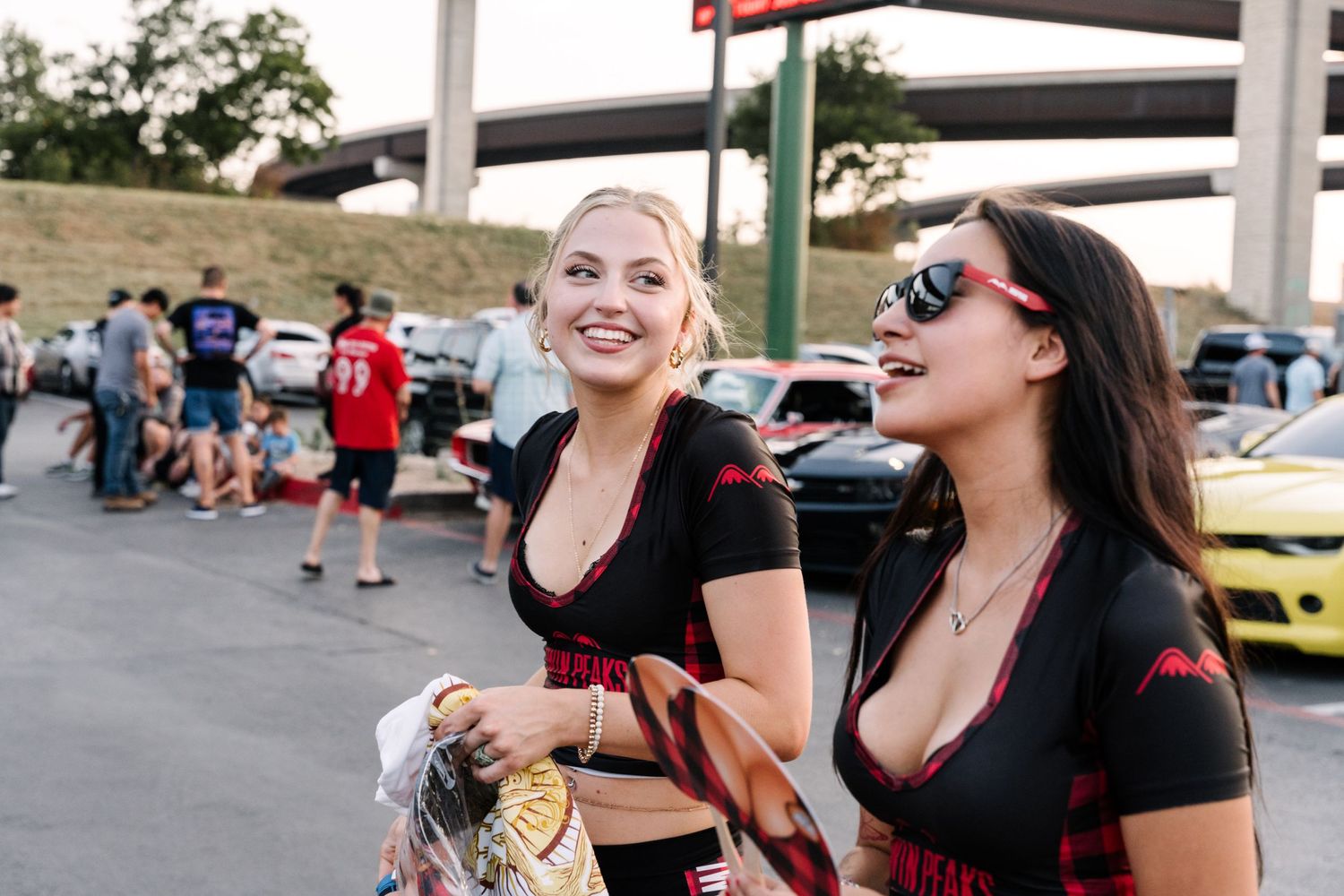 Two friends sharing a laugh at a car meetup event in a parking lot.
