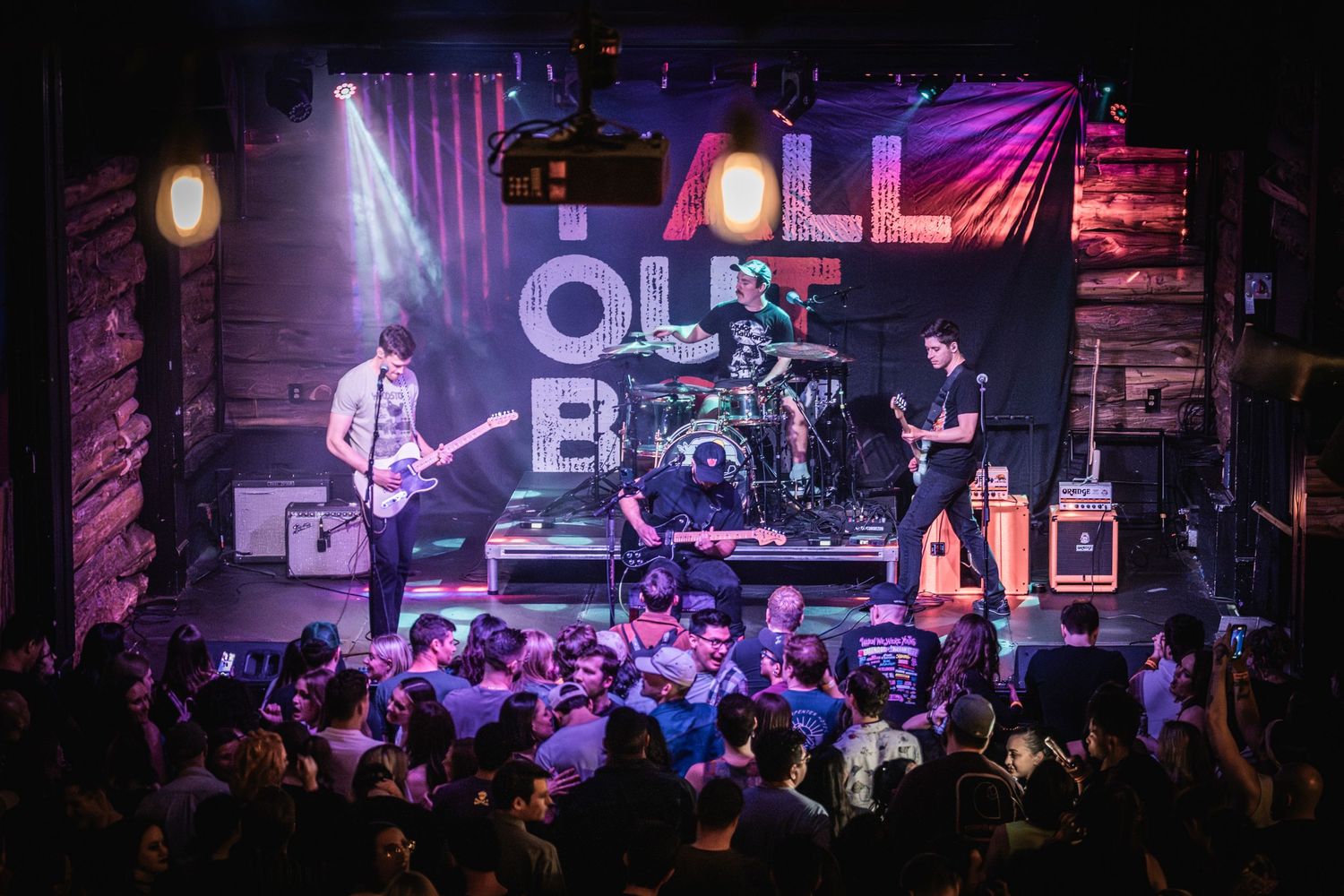 Rock band performing on stage at a crowded music venue with purple lighting.