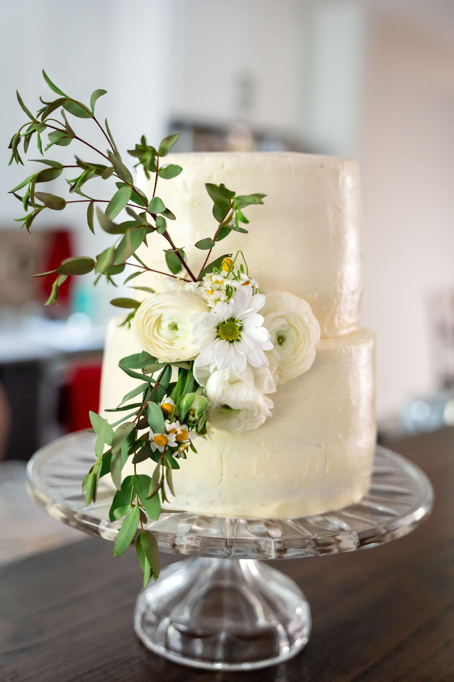 Simple white wedding cake decorated with fresh white flowers and greenery on crystal cake stand.