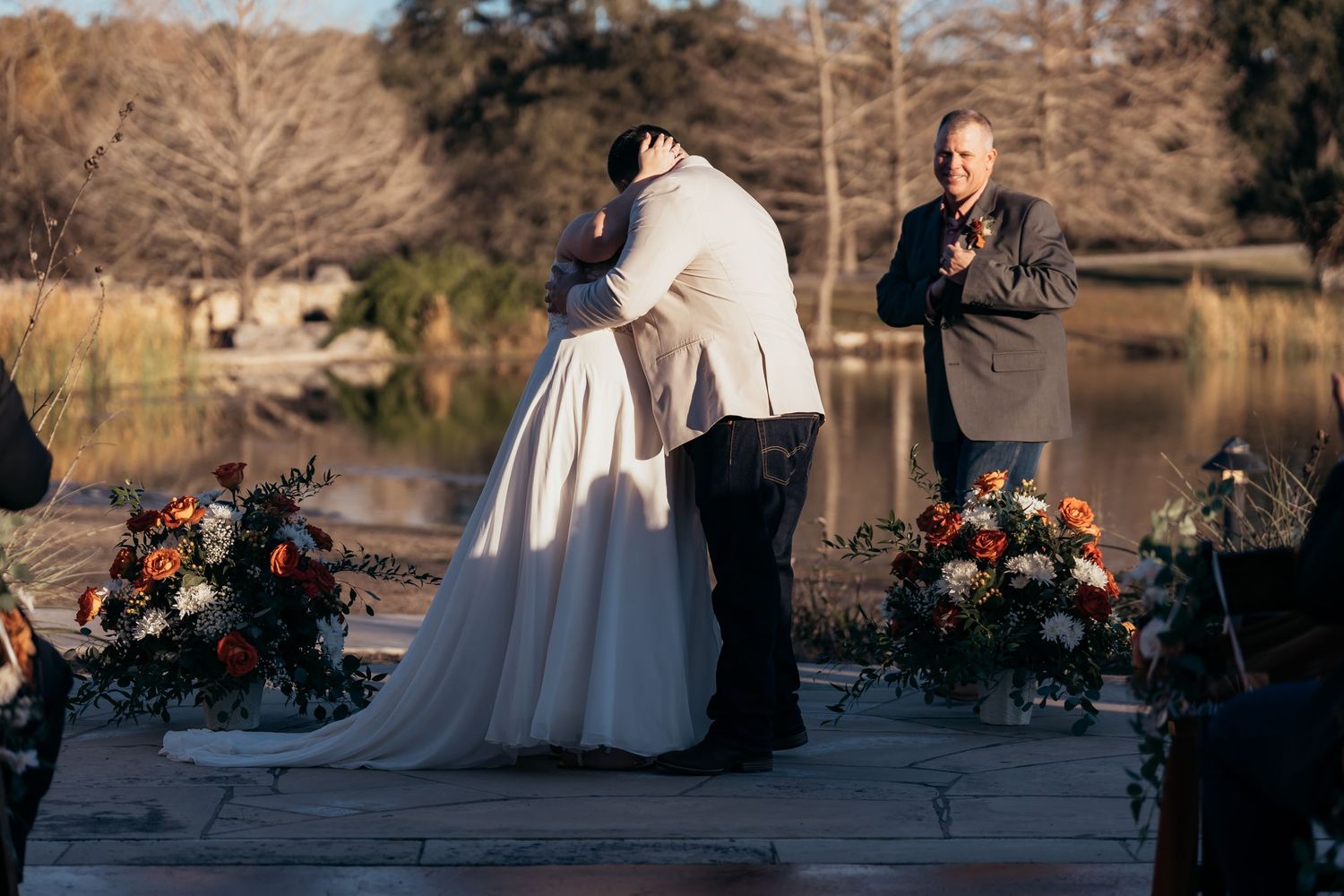 A wedding ceremony takes place by a lake with floral arrangements as the couple embraces at sunset.