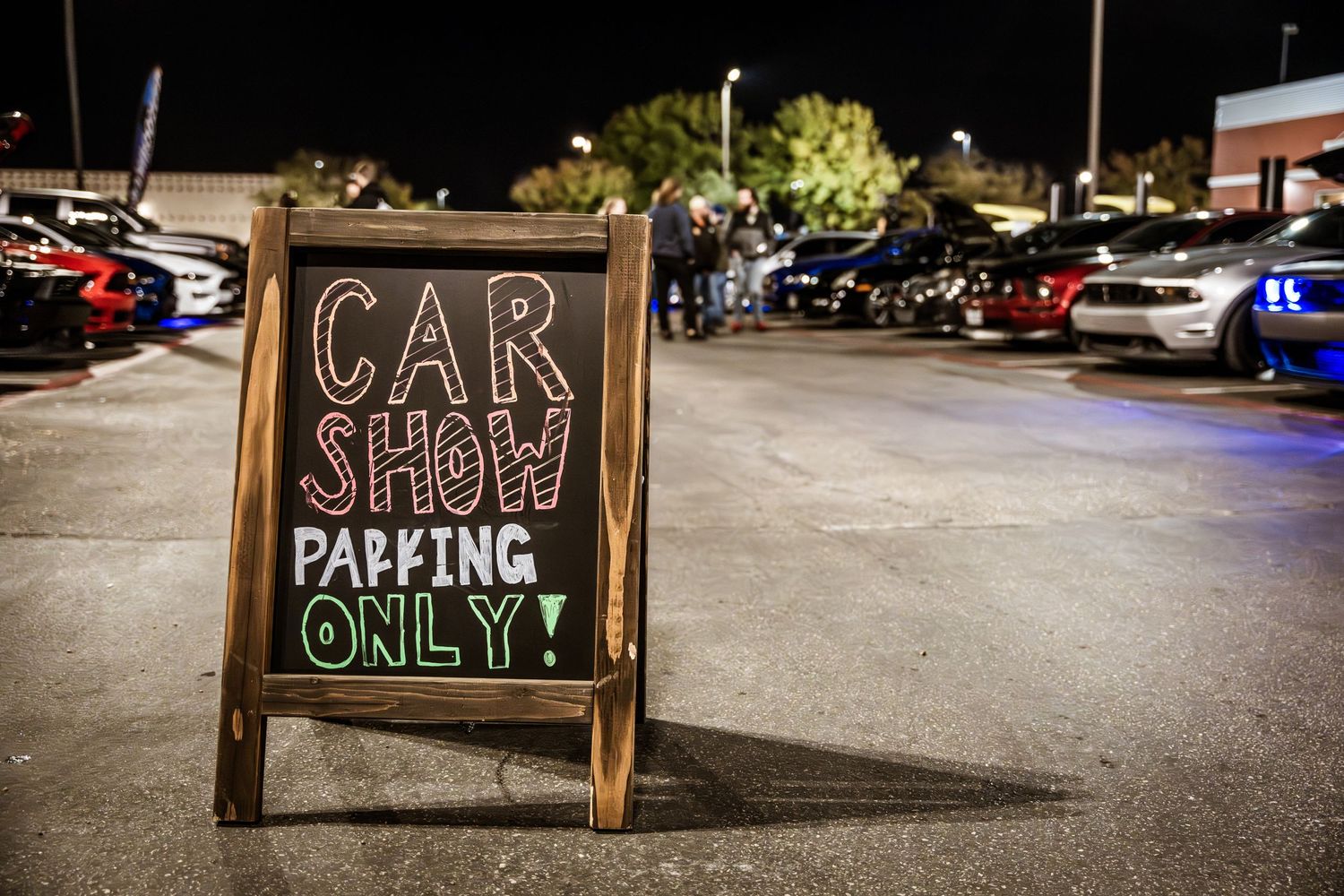 A chalkboard sign reading 'Car Show Parking Only' stands in a nighttime parking lot.