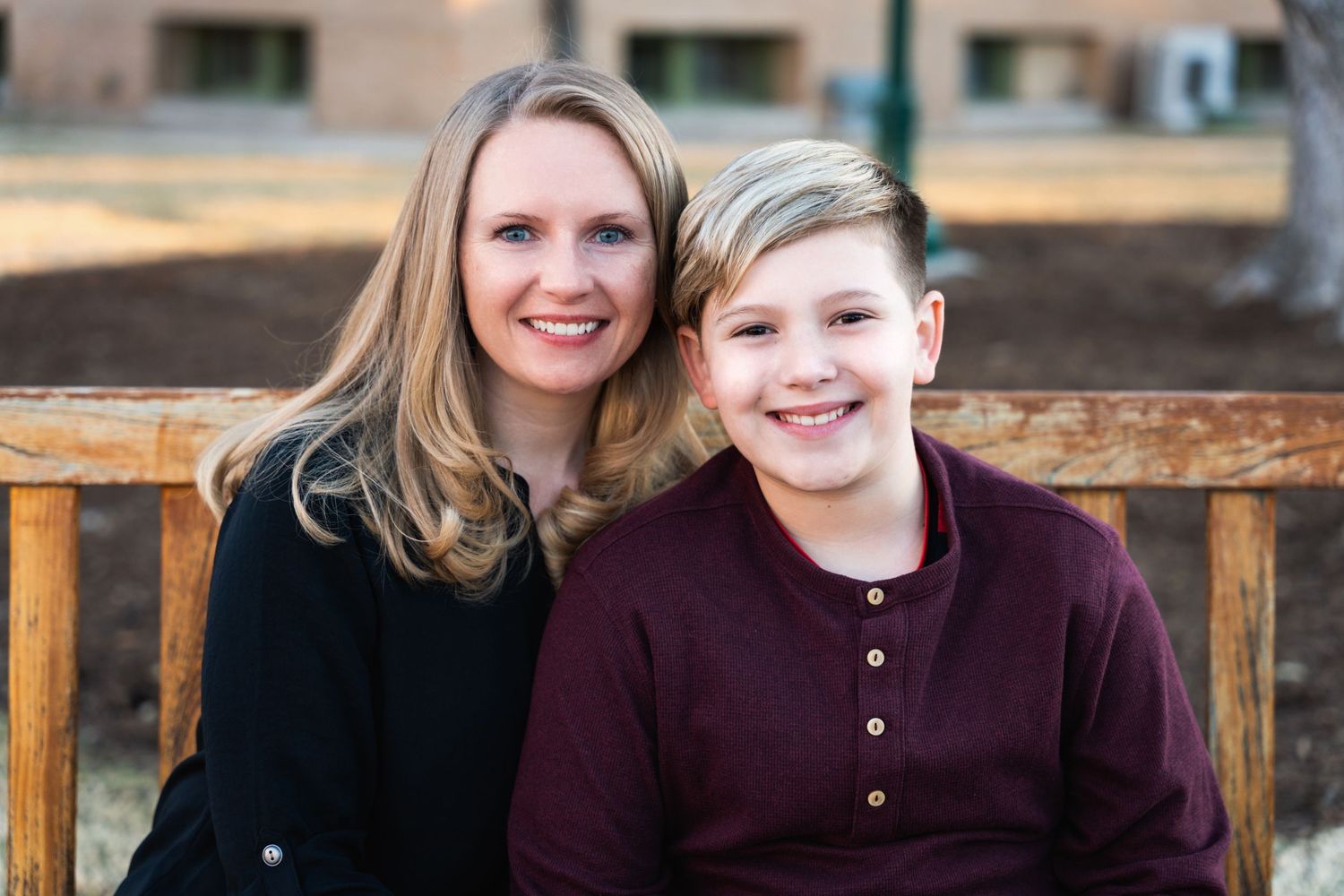 Portrait of two people in black and burgundy sitting on wooden bench outdoors.